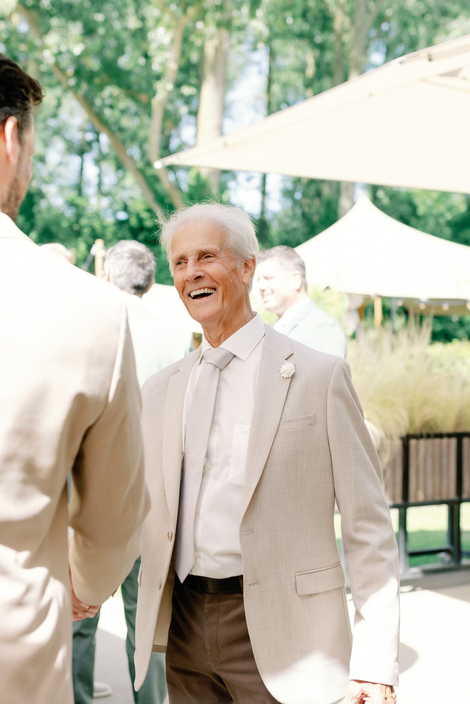 Wedding party members in light beige suits share a candid laughing moment outdoors during a celebration.