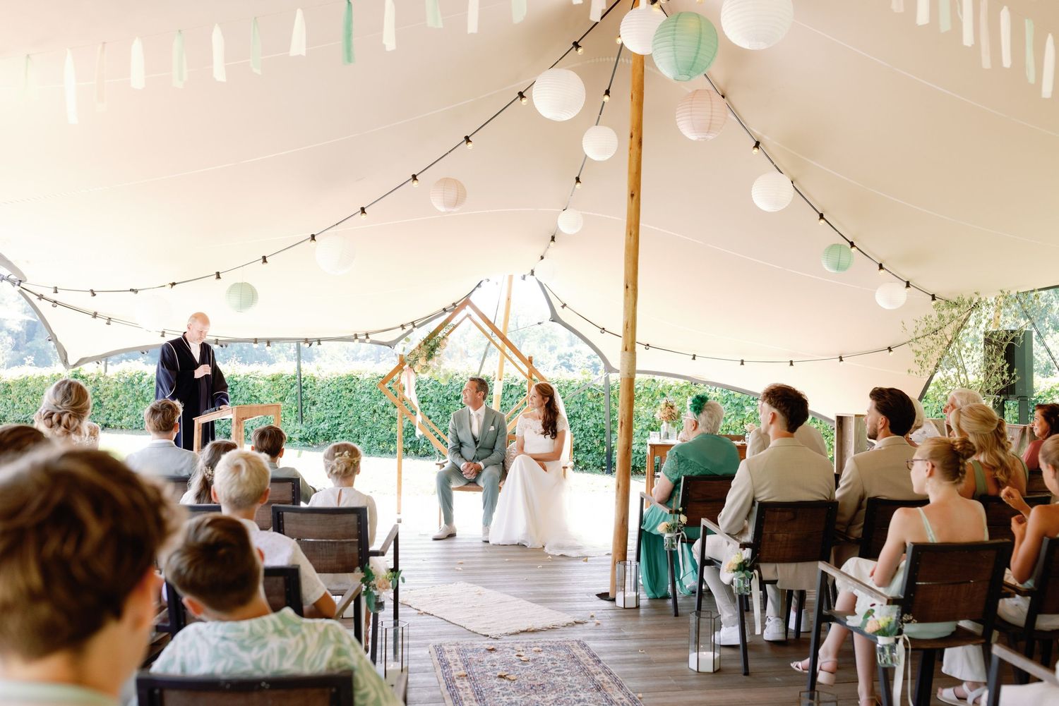 Wedding guests sit under an elegant white tent decorated with string lights during an outdoor reception celebration.
