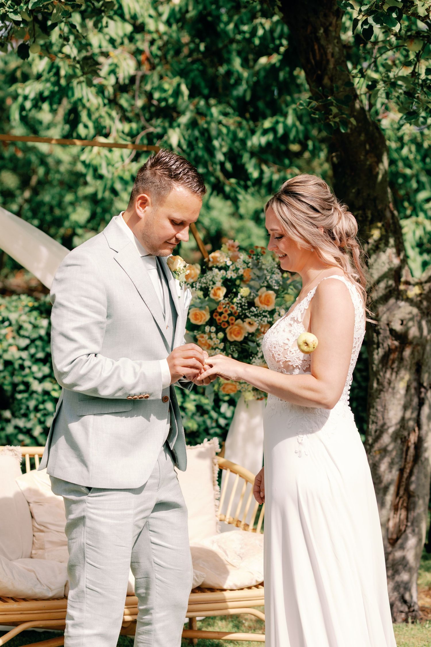 A couple in white attire exchanges rings during an outdoor garden wedding ceremony.