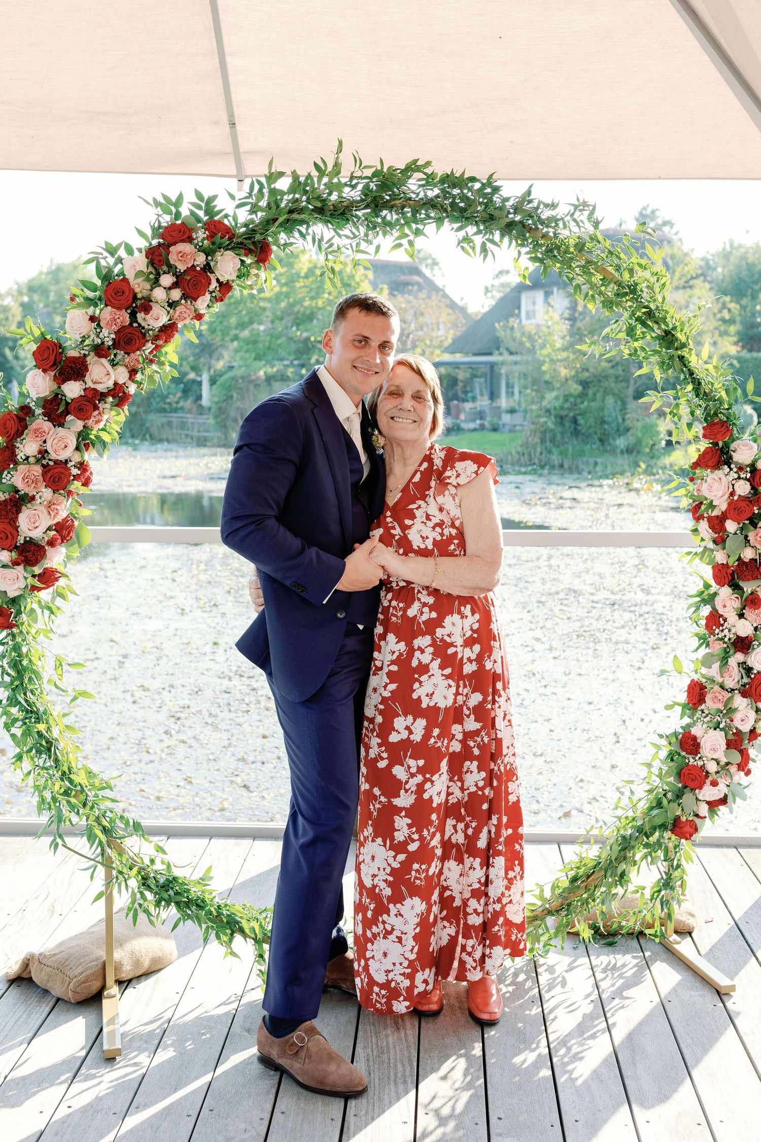 Couple poses under floral arch with red and white flowers on waterfront deck during romantic photoshoot.