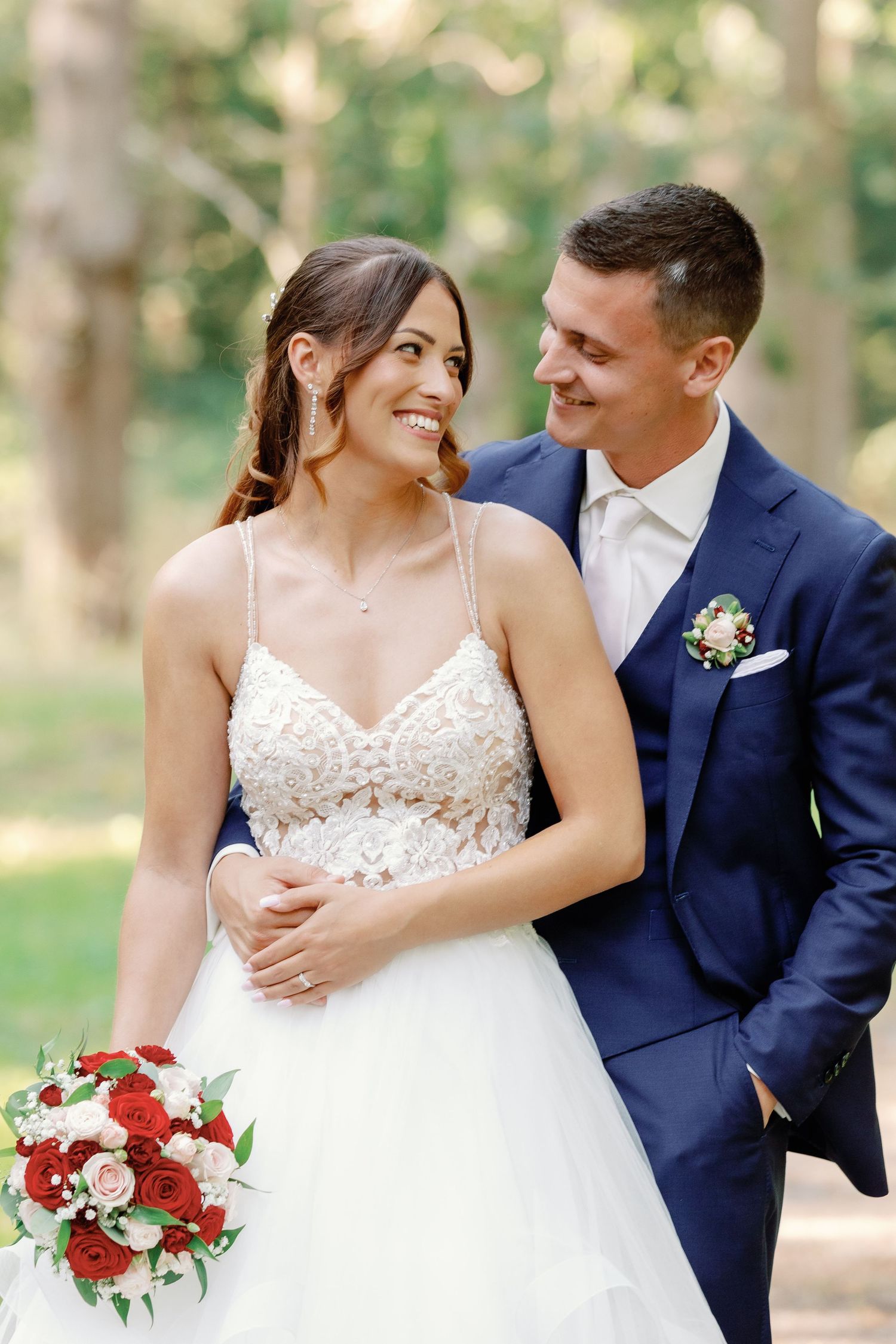 Wedding couple in navy suit and white dress holding red bouquet in outdoor garden setting.