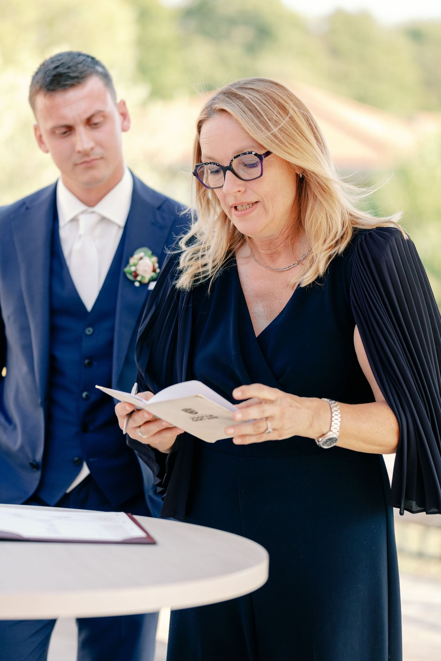 A wedding officiant in a navy dress reads from notes during an outdoor ceremony.