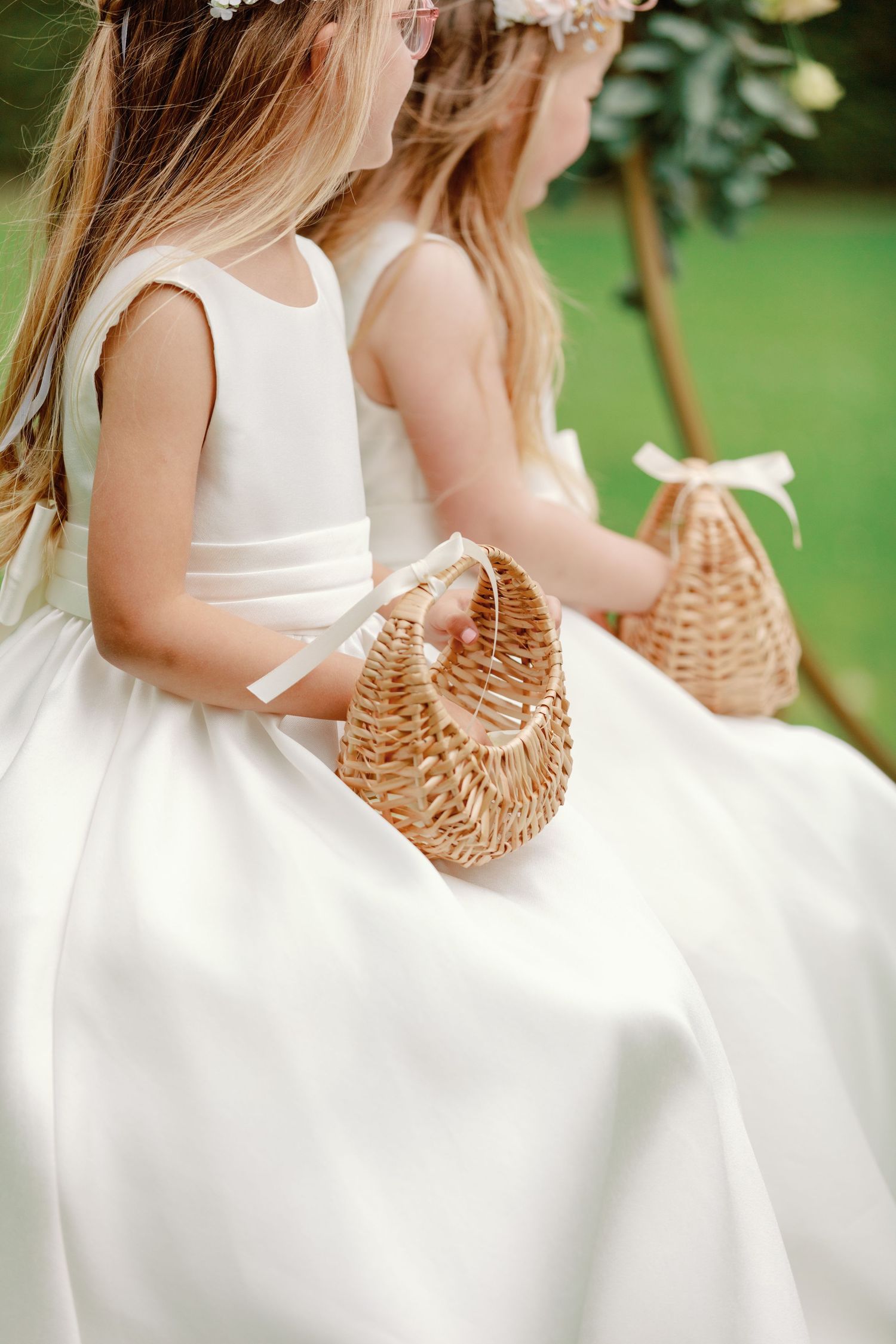A row of flower girls in white dresses holding woven basket purses at an outdoor wedding ceremony.