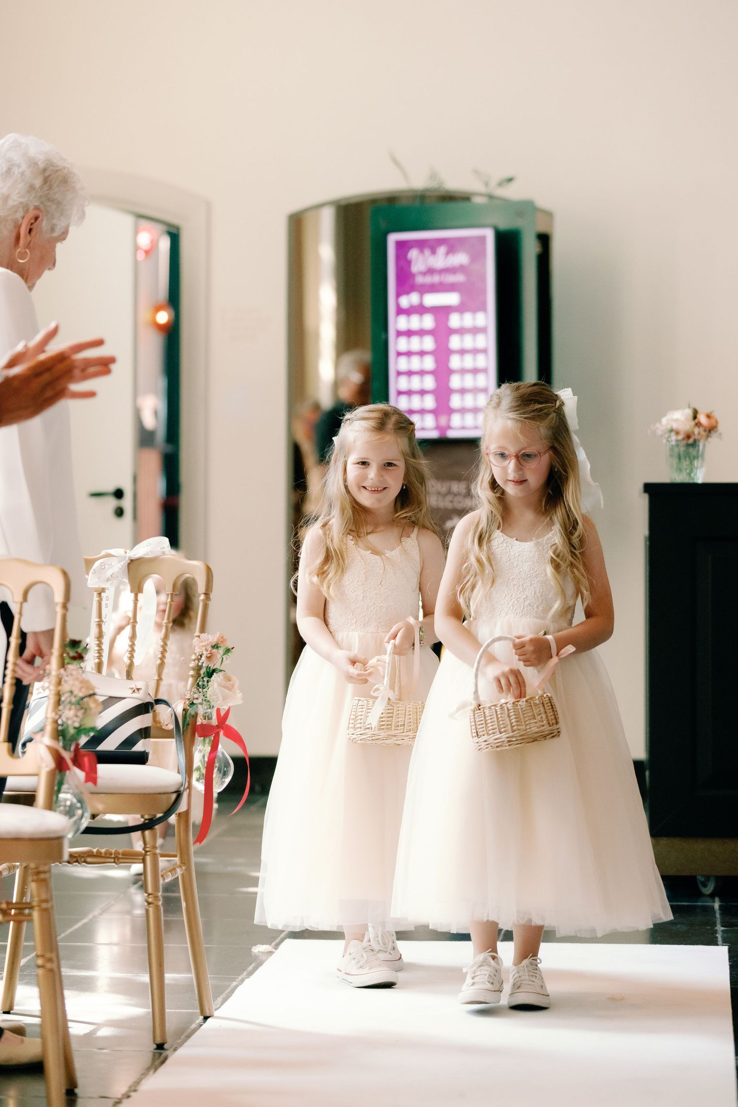 Young flower girls in white dresses holding woven baskets walk together in a bridal preparation room.