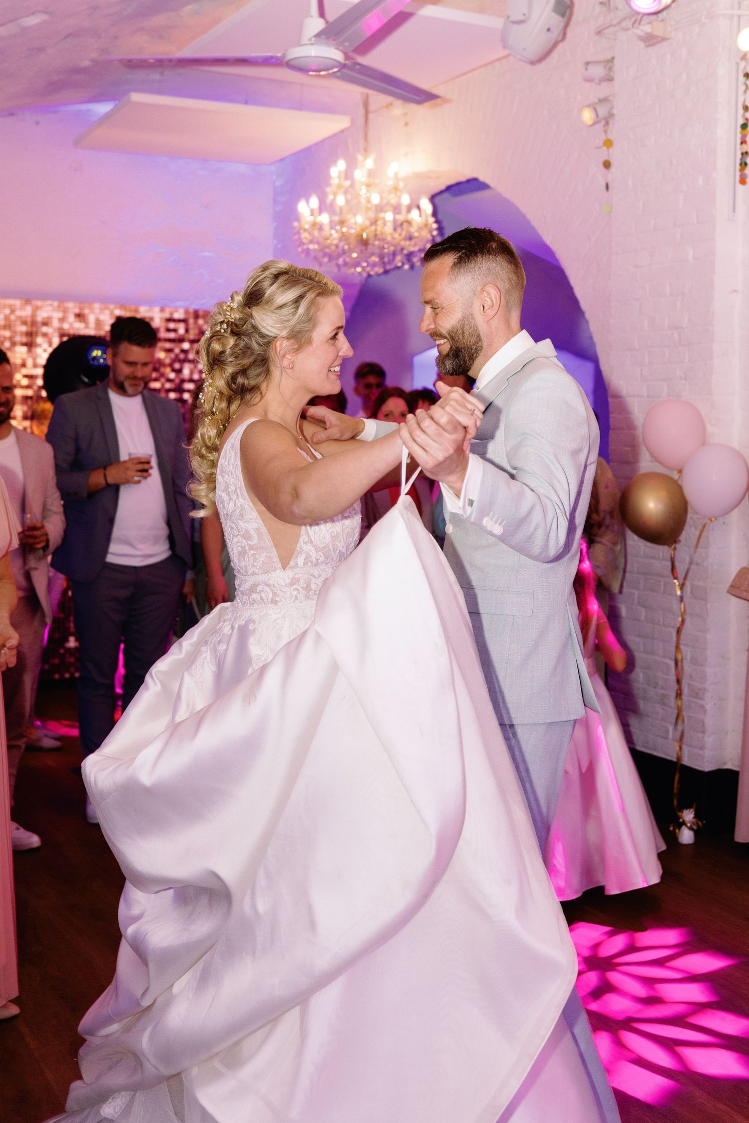 A couple shares a romantic dance moment at a wedding reception with pink lighting and elegant chandeliers in the background.