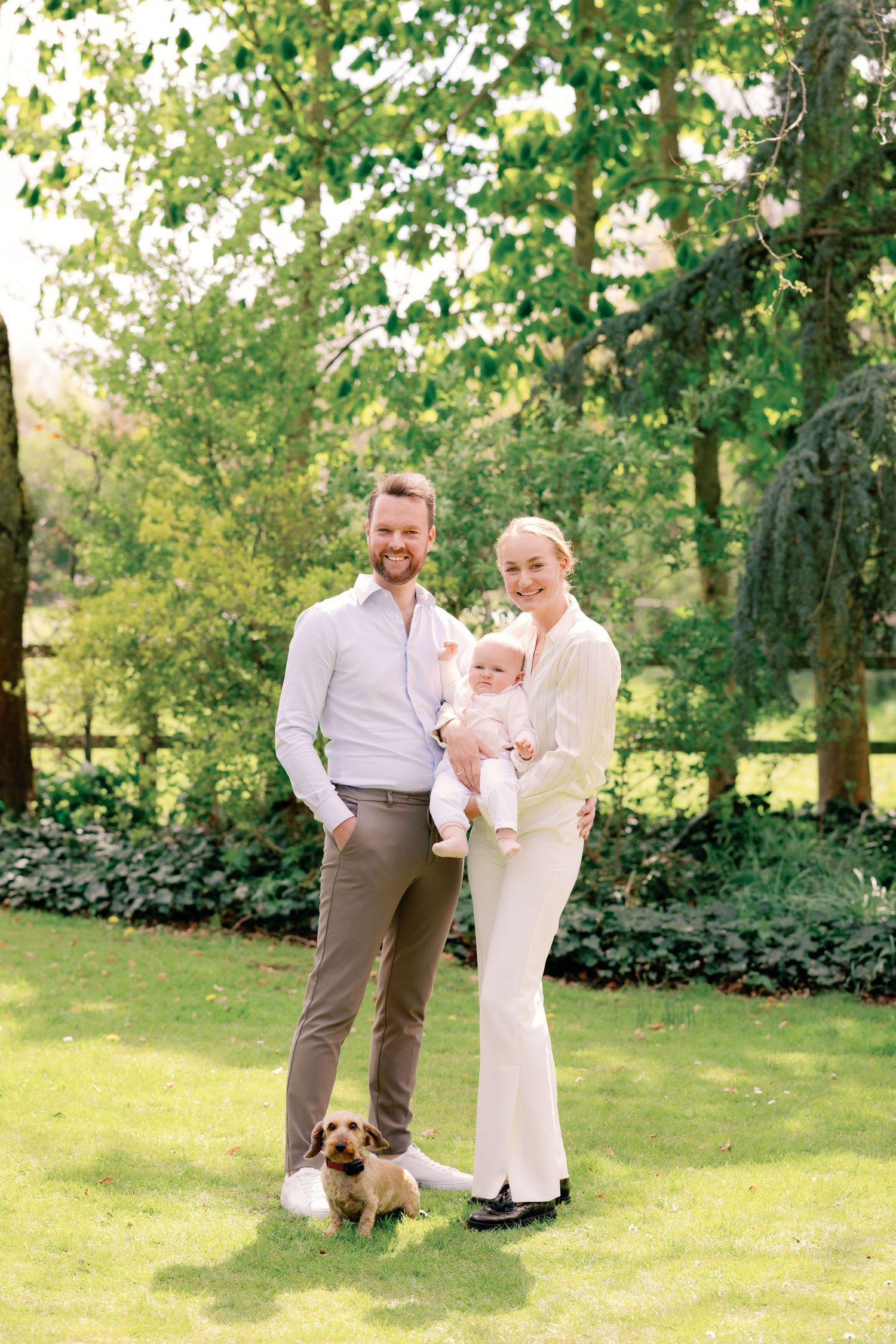 A happy family with a baby and small dog posing in a lush green garden on a sunny day.