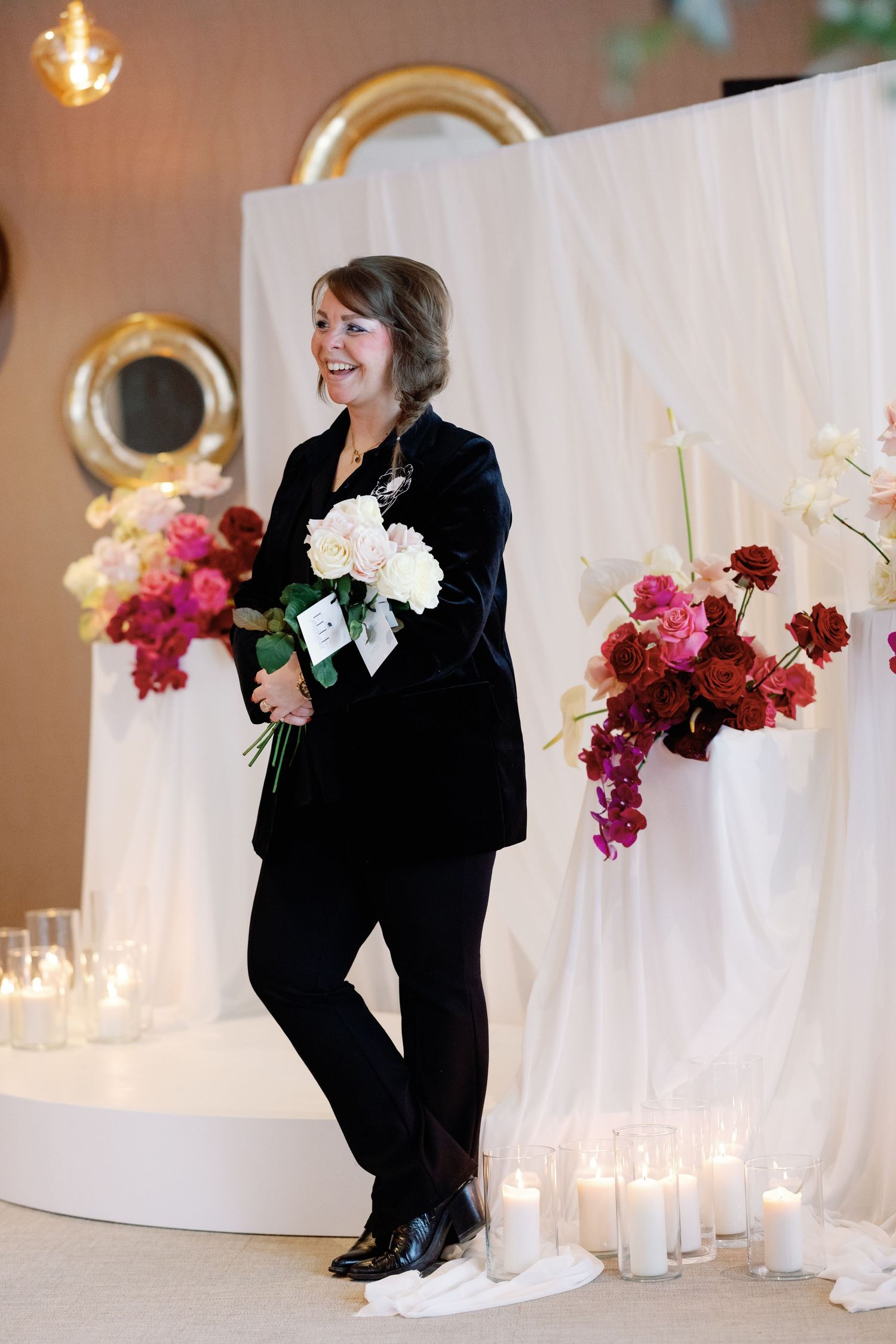 A person in black attire arranges colorful flowers against a white backdrop with glowing candles.