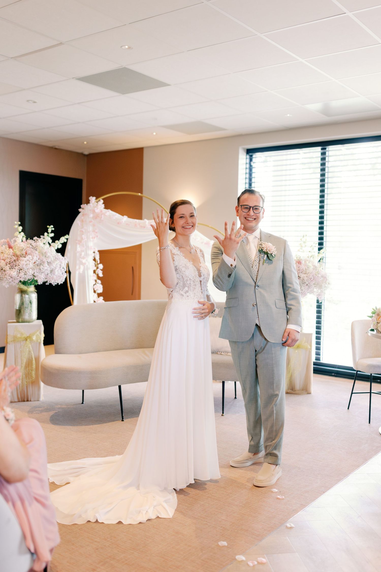 Bride and groom showing wedding rings at elegant indoor ceremony with floral arch and neutral decor.