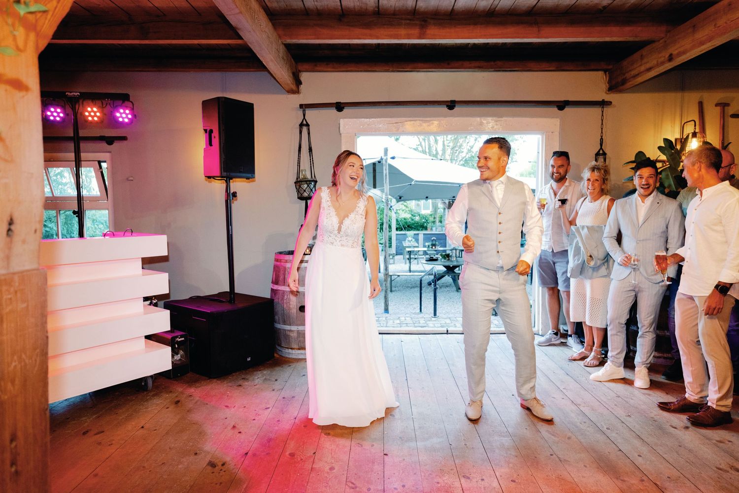 A couple shares their first dance at a rustic indoor venue with wooden beams and soft pink lighting.