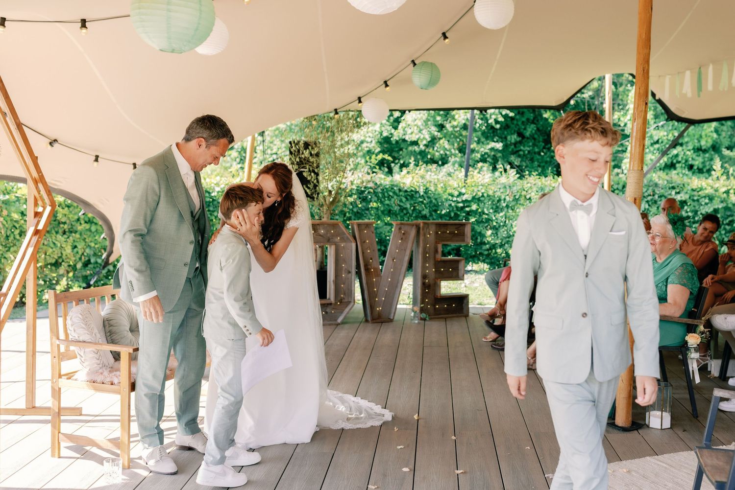 Wedding guests in white attire mingle on an outdoor covered patio decorated with string lights and greenery.