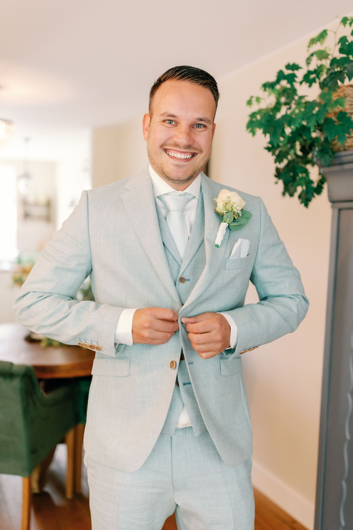A person wearing a light blue suit adjusts their tie while standing in a bright indoor space with greenery in the background.