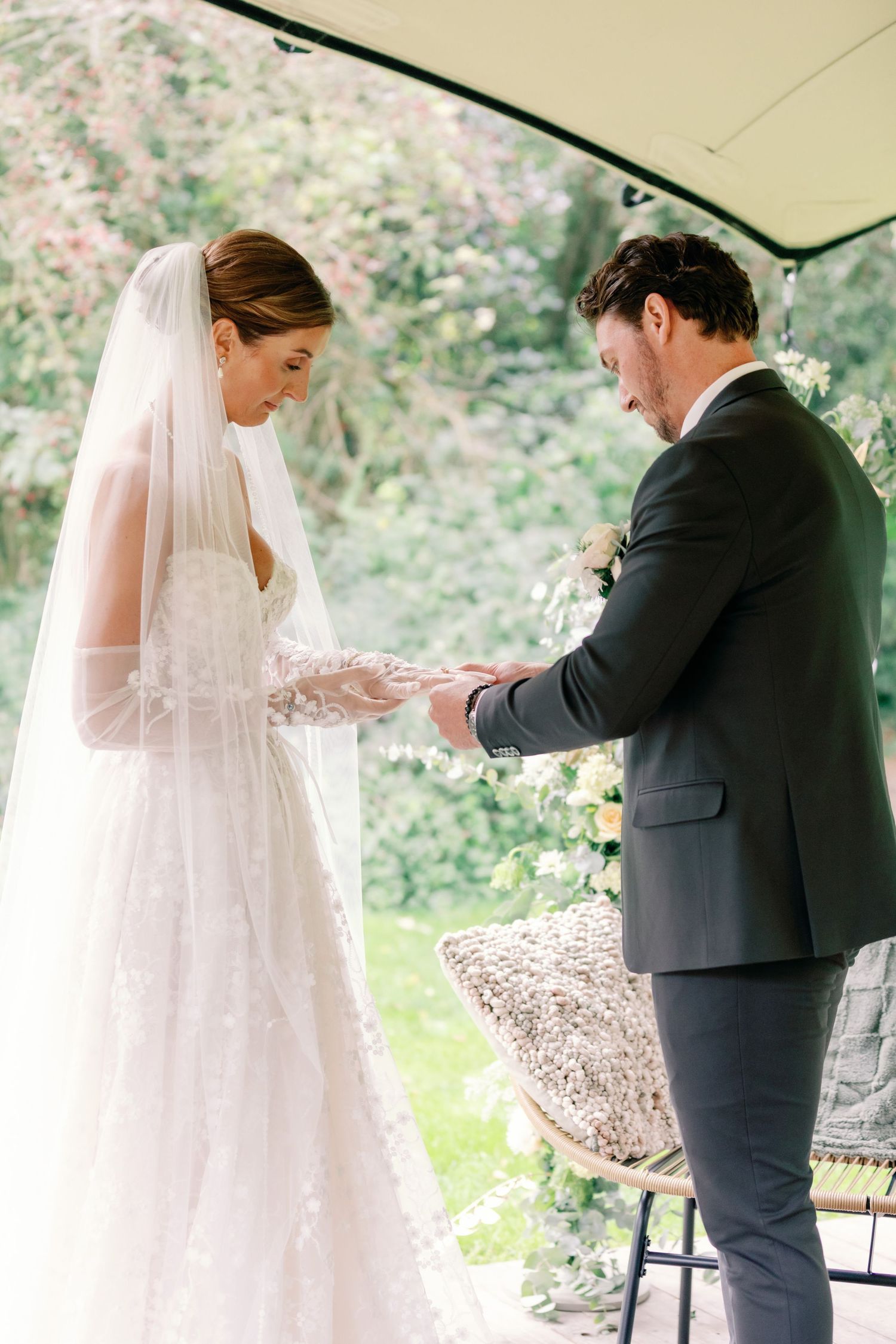 A tender moment at a wedding ceremony as a couple exchanges rings under an outdoor canopy with greenery in the background.