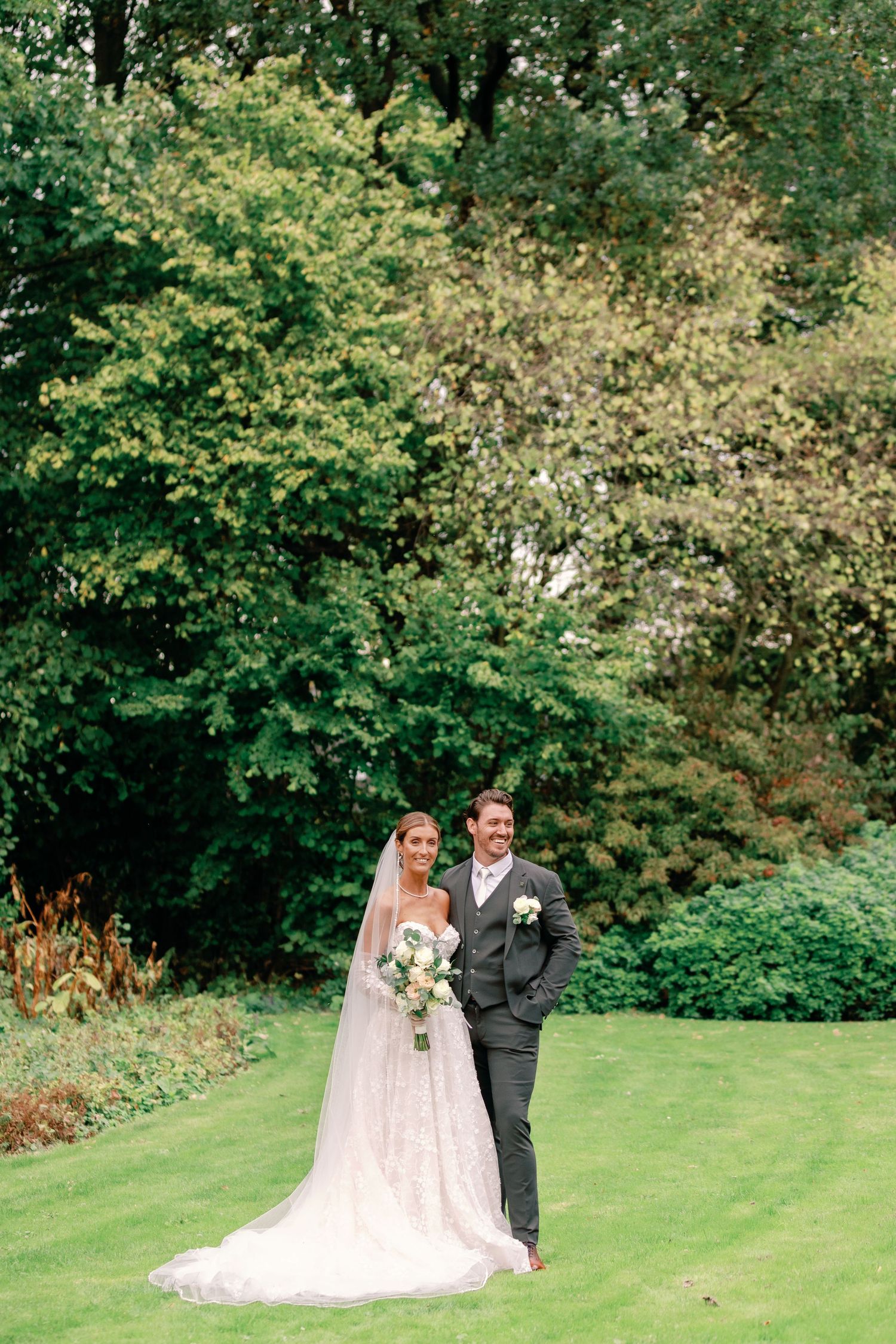 A couple in wedding attire share a tender moment on a lush green hillside surrounded by trees.