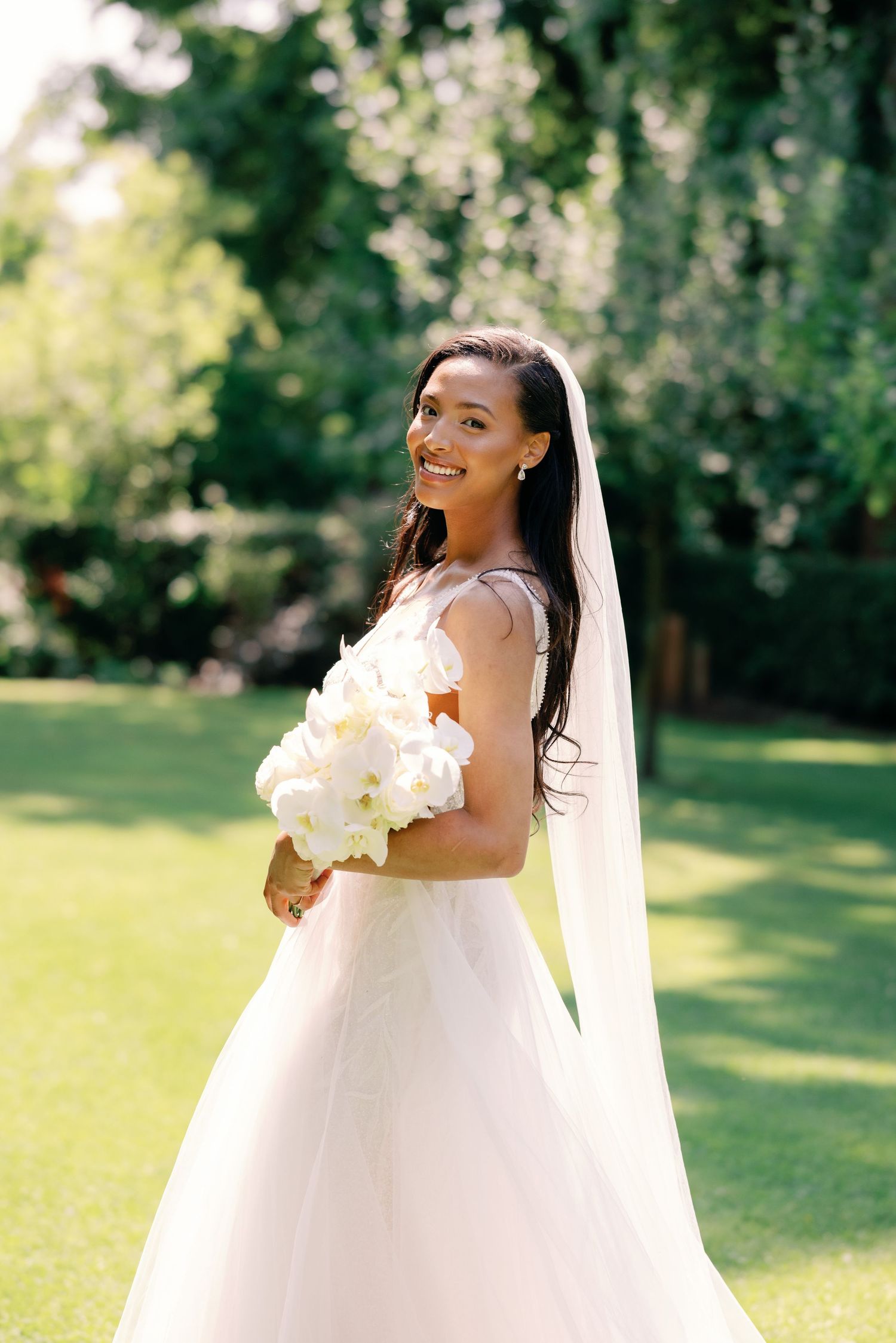 A bride in a white ballgown wedding dress holds a bouquet of white flowers in a sunny garden setting.