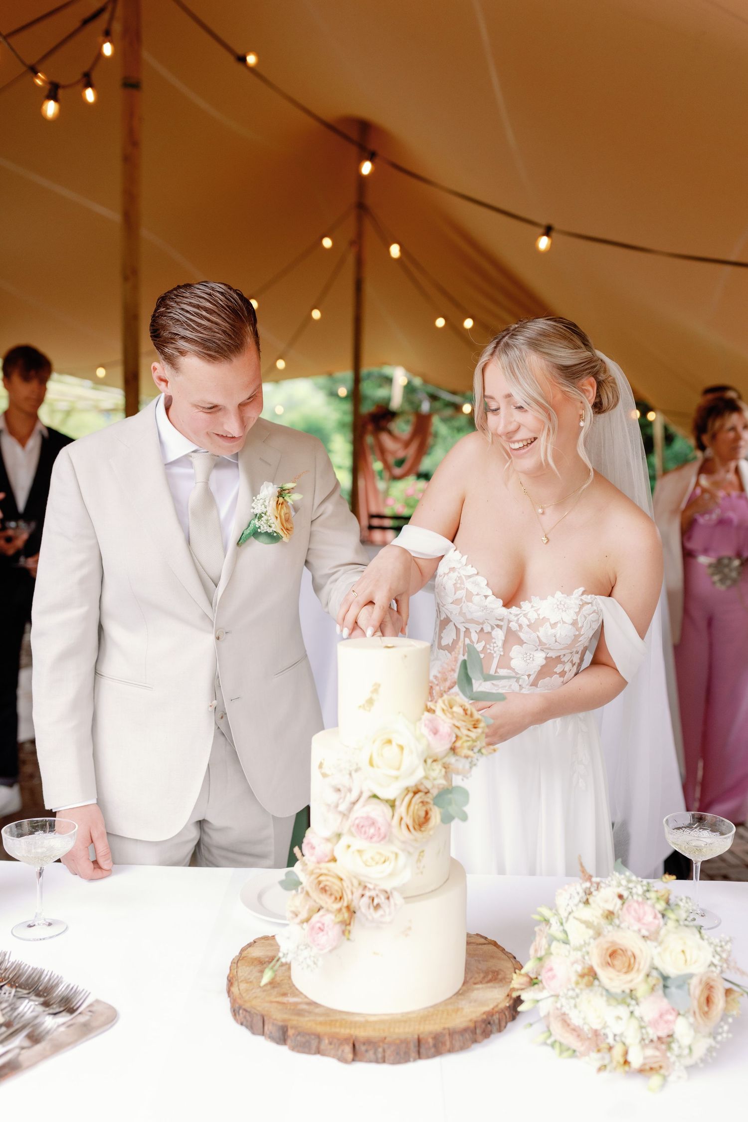 Newlyweds cut their elegant white wedding cake decorated with peach flowers on a rustic wood slice stand under a tent.