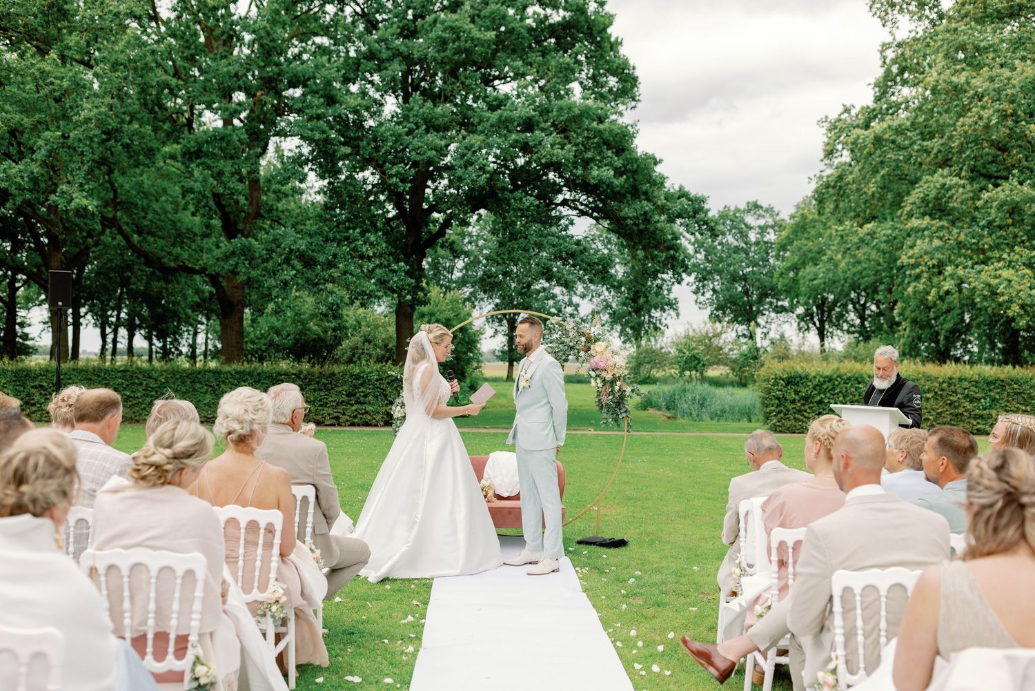 Outdoor wedding ceremony on a lush green lawn with white chairs and tall trees creating a natural backdrop.