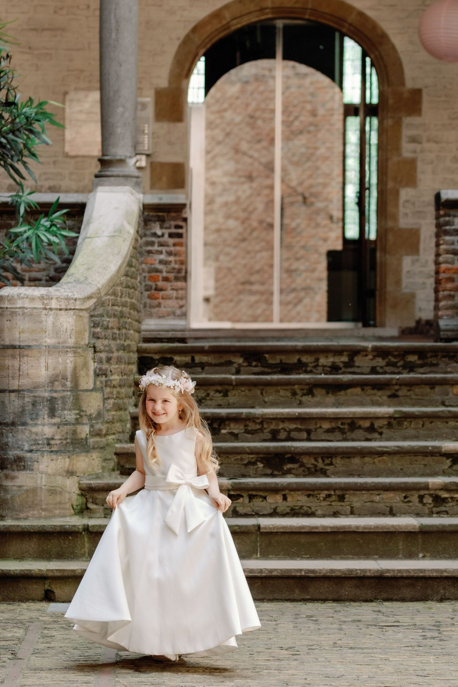 A young flower girl in a white dress twirls and plays on stone steps in a historic courtyard setting.