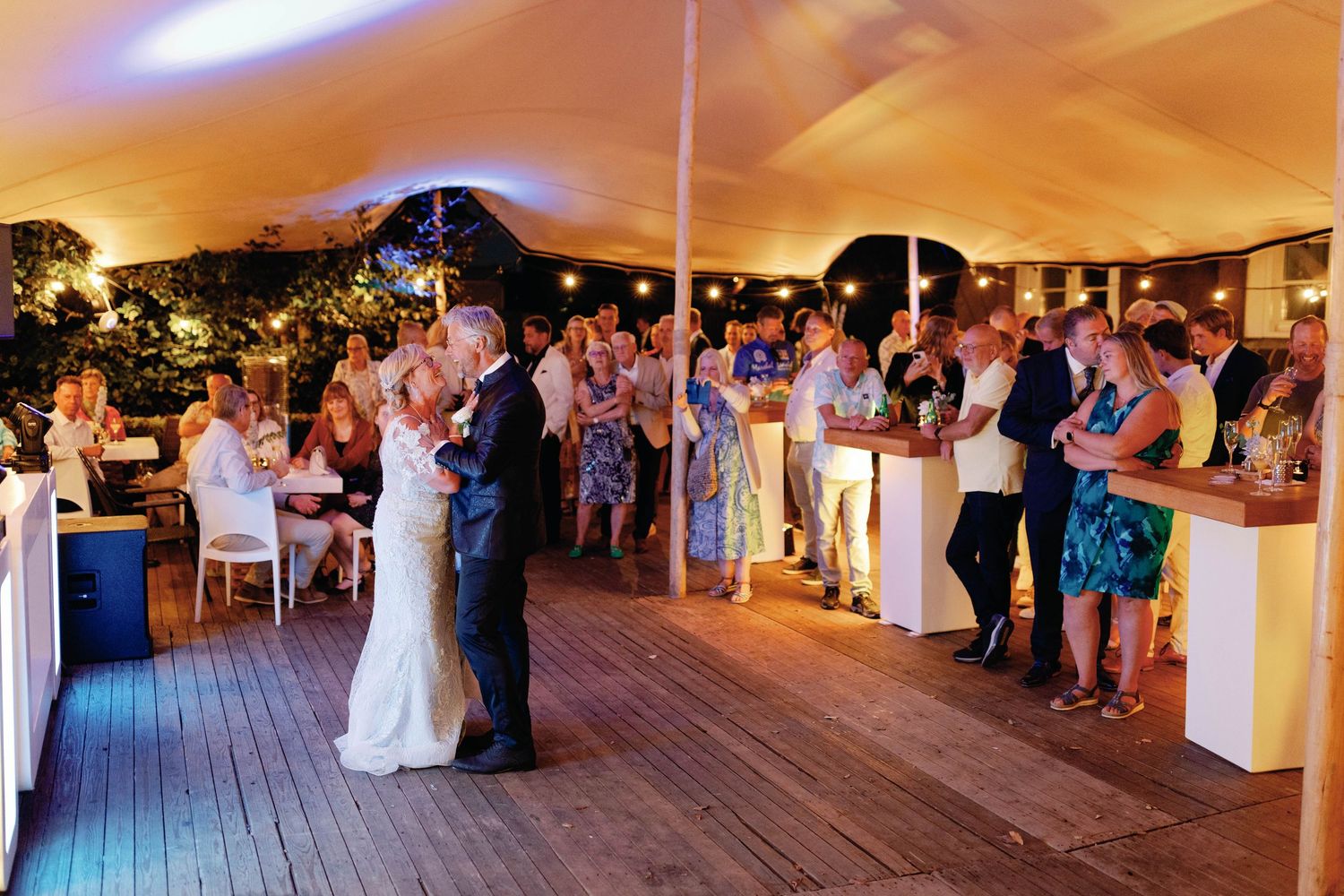 Large wedding reception celebration under warm-lit tent with guests seated at tables.