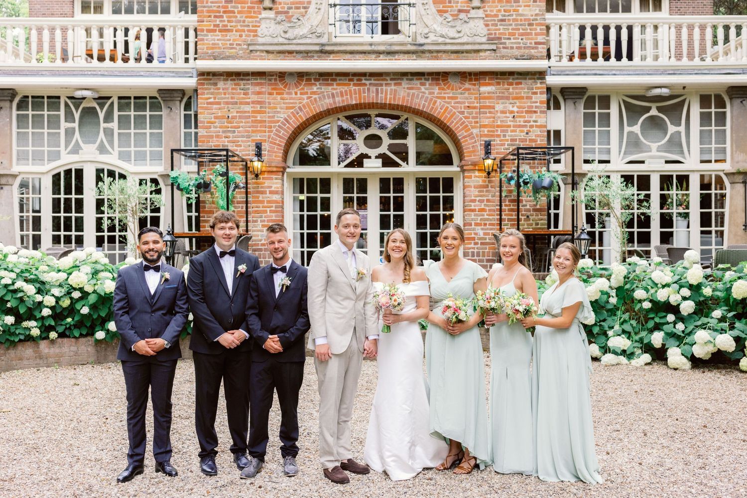 Wedding party posing in front of a historic brick building with arched windows and blooming hydrangeas.