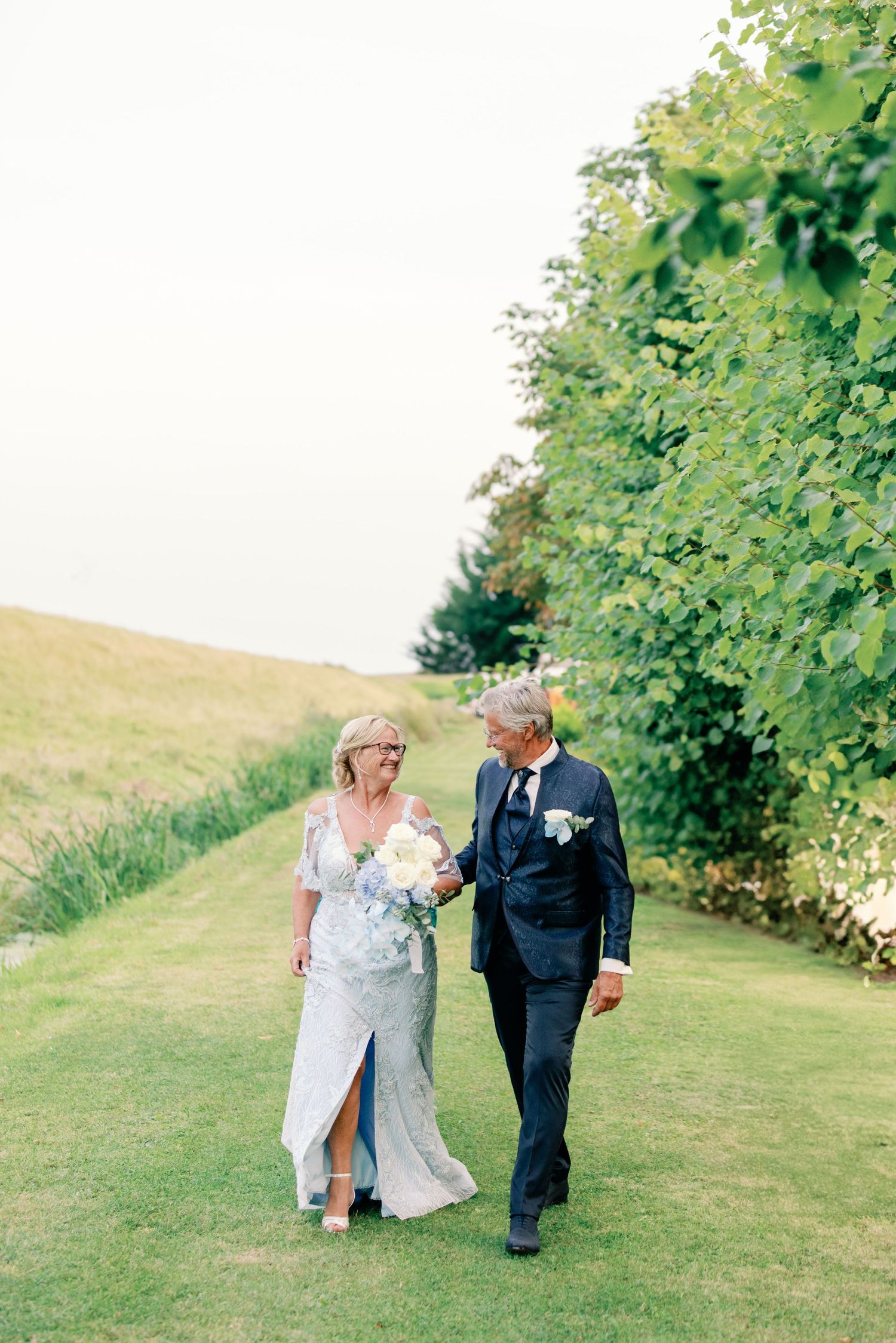 Couple walks together on grassy hillside during outdoor wedding celebration.