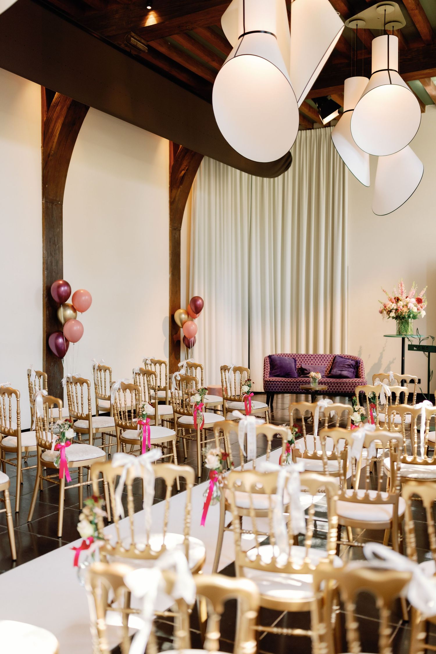 Elegant wedding reception venue with round tables, gold chairs, white paper lanterns, and pink balloon accents.