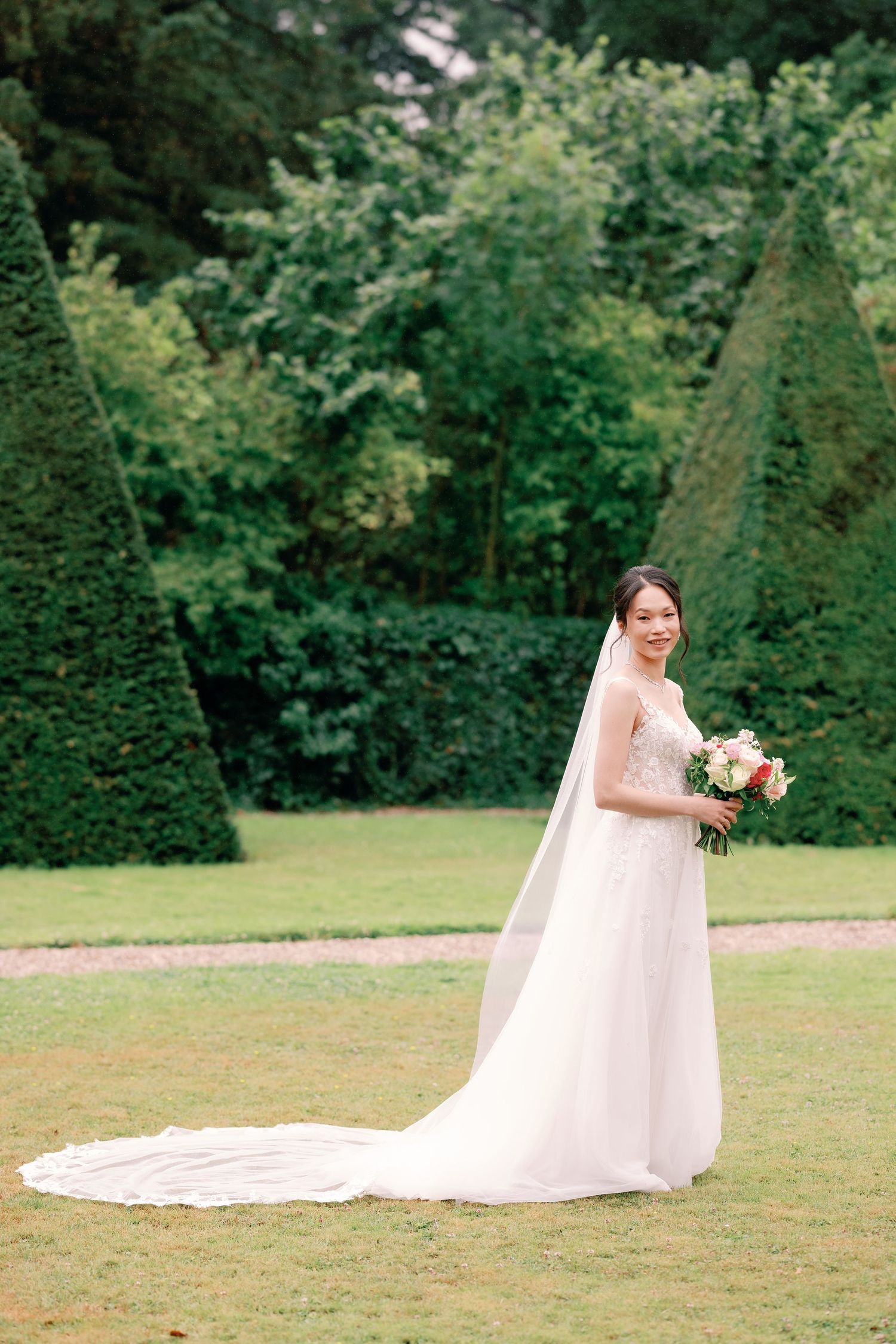 A bride in a flowing white wedding gown with long train poses gracefully on a green lawn surrounded by trees.