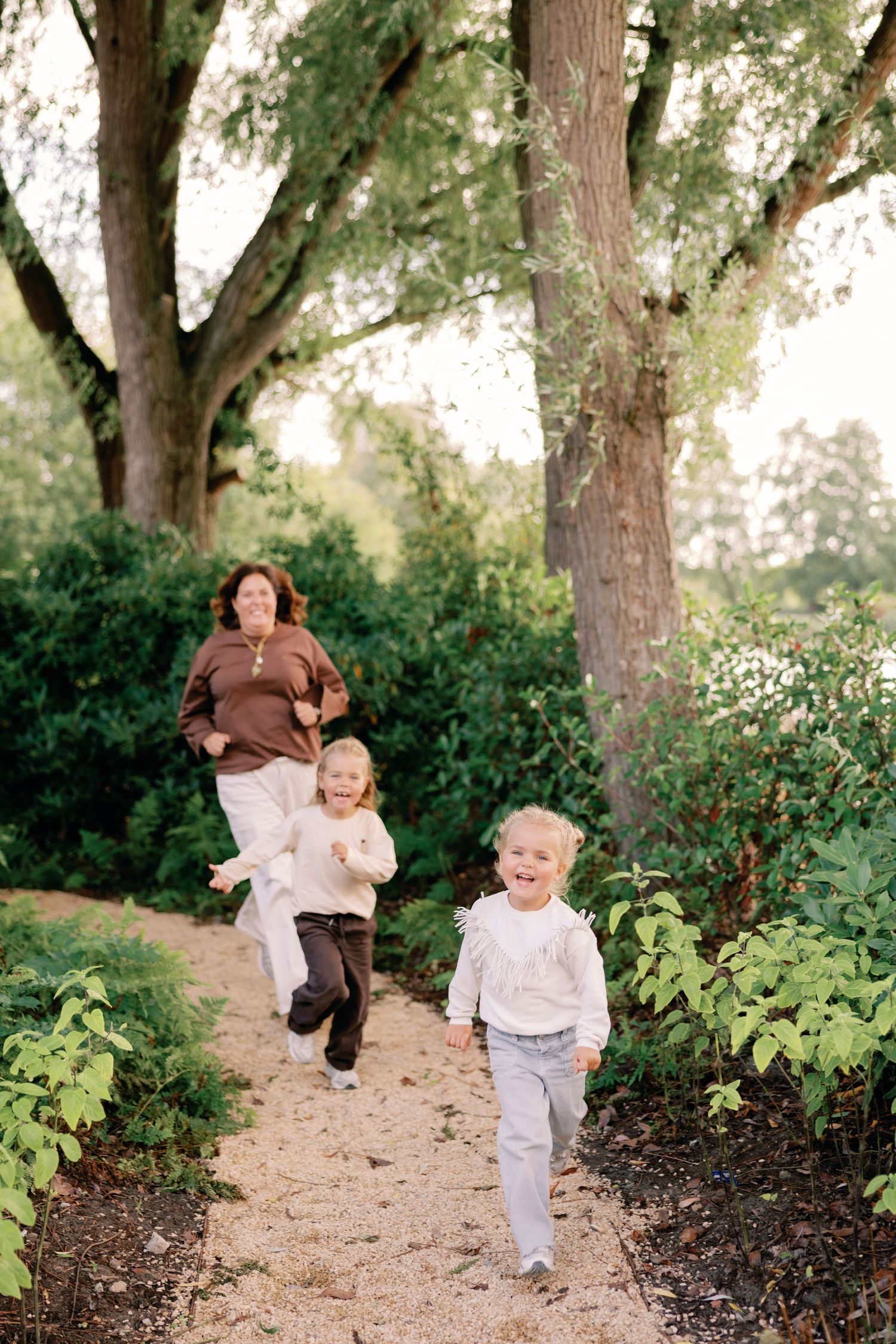 Two laughing young children run down a gravel path through lush green trees while a smiling woman follows behind.