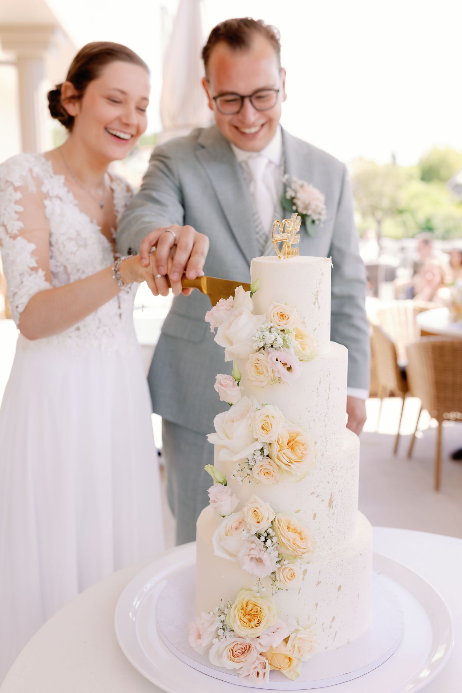 Bride and groom smiling while cutting a elegant four-tier white wedding cake adorned with fresh roses.