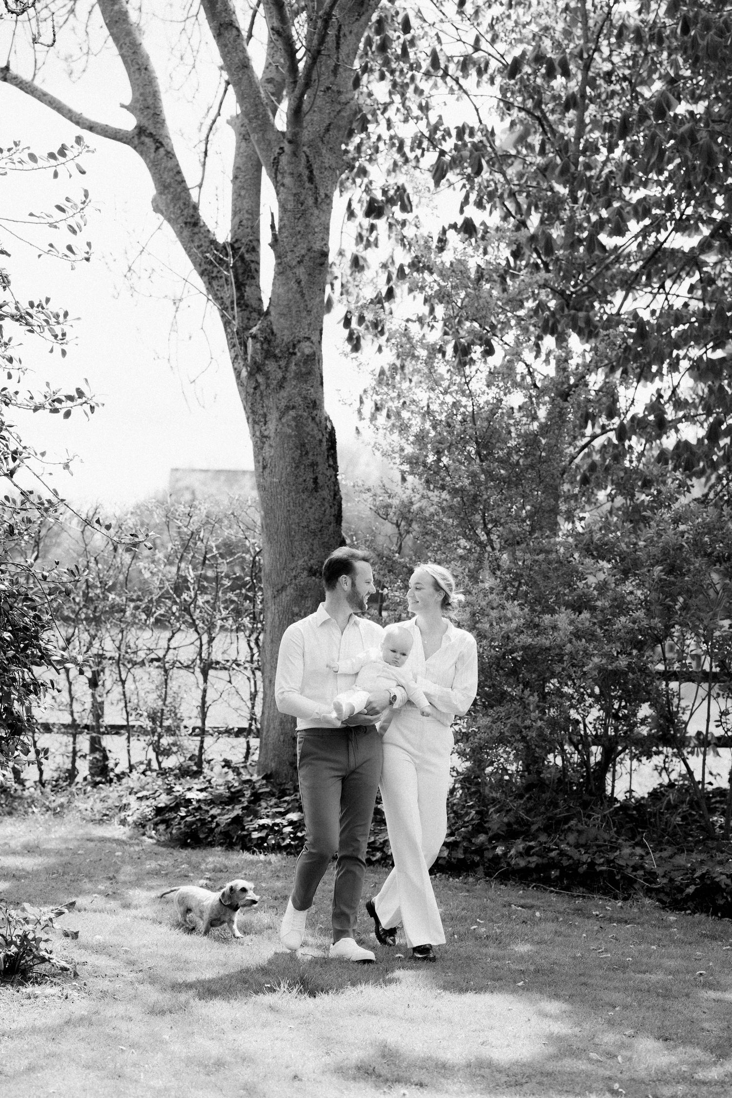 Black and white photo of a couple holding a baby outdoors near a large tree with a small dog nearby.