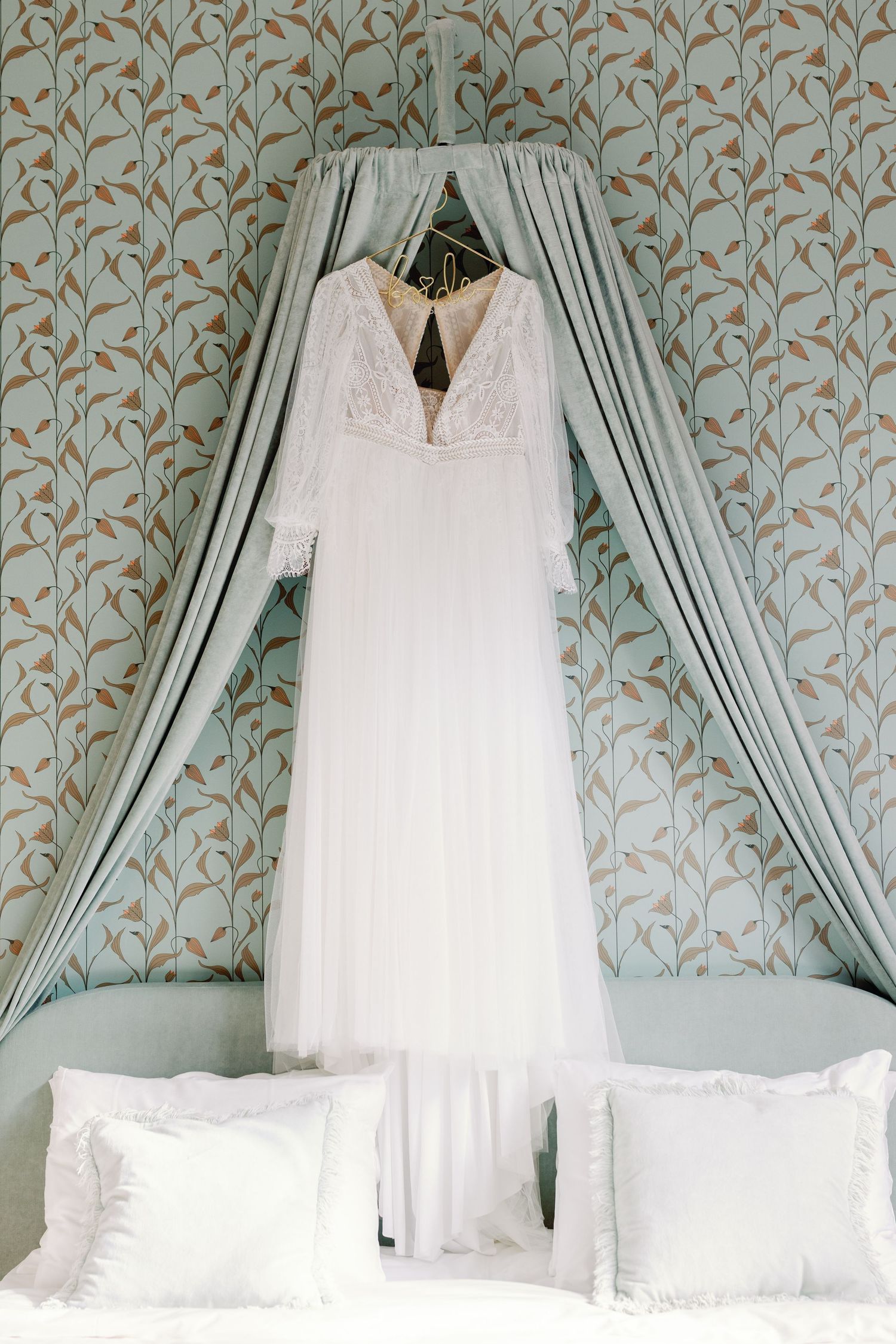 A white lace wedding dress hangs elegantly against a blue floral wallpaper background in a bridal preparation room.