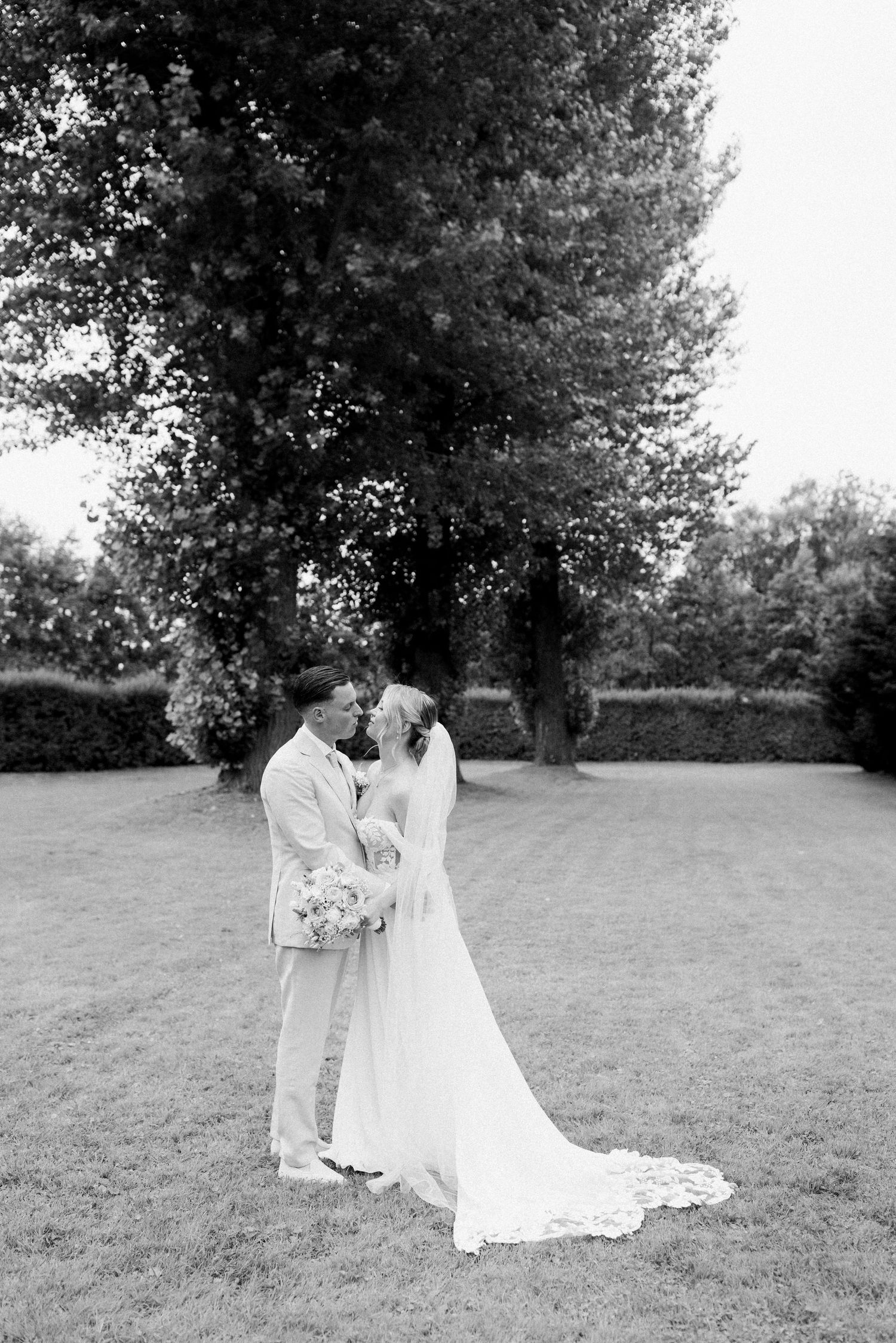 A black and white wedding photograph of a couple embracing on a lawn with tall trees in the background.