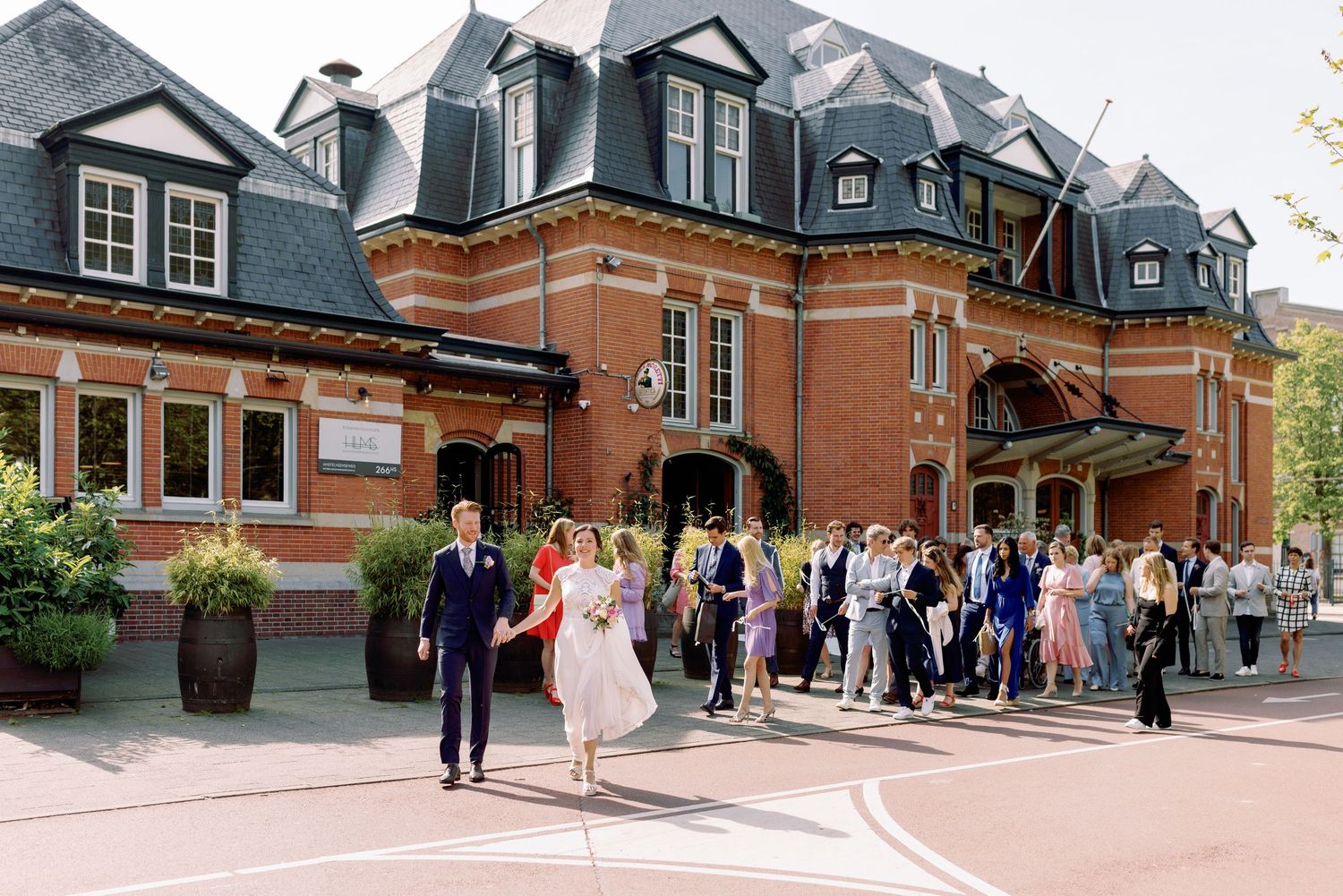Panoramic view of people gathering outside a historic brick building with dormers and dark slate roof.
