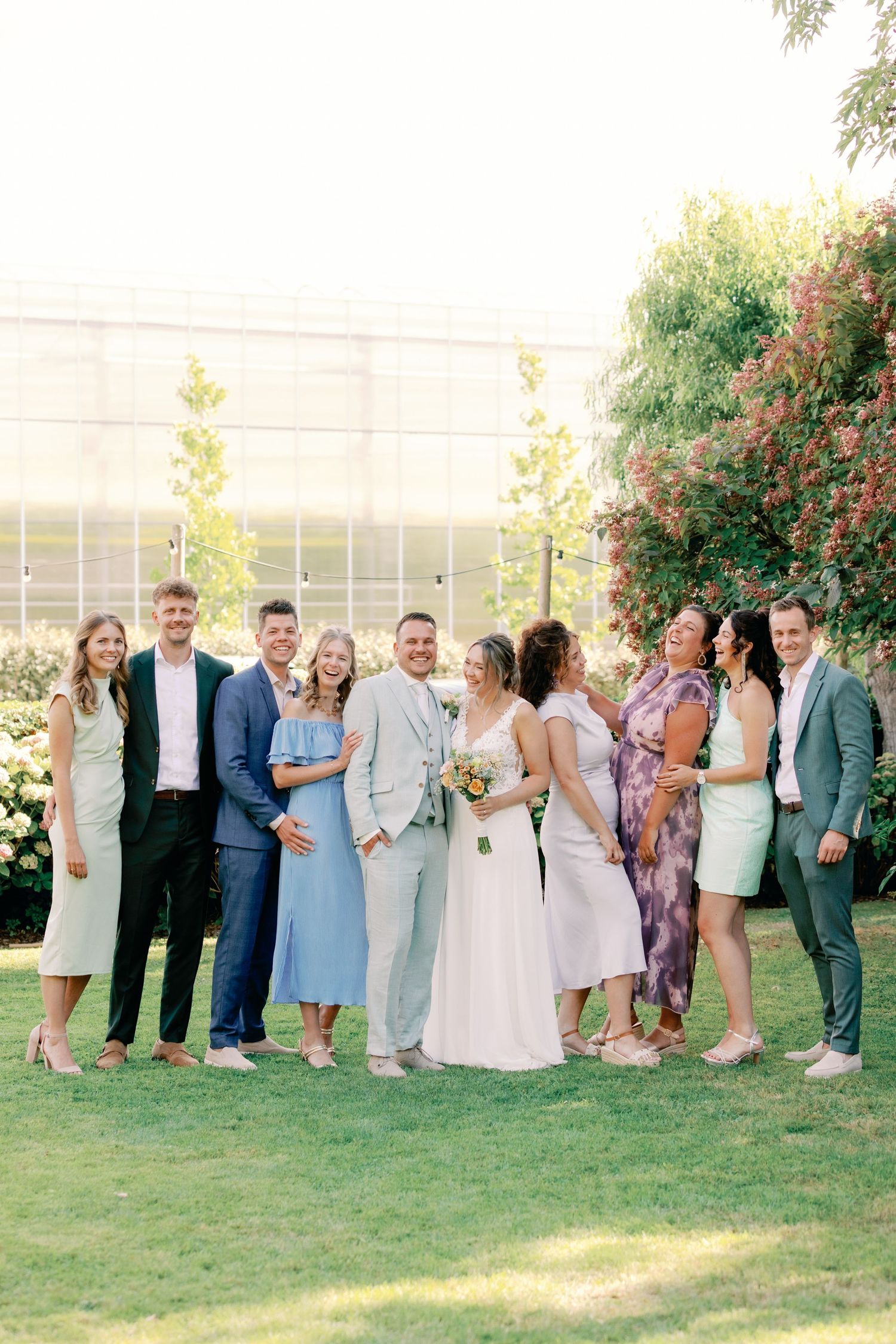 Wedding party stands in line on green lawn with flowering bushes and building in background during outdoor celebration.