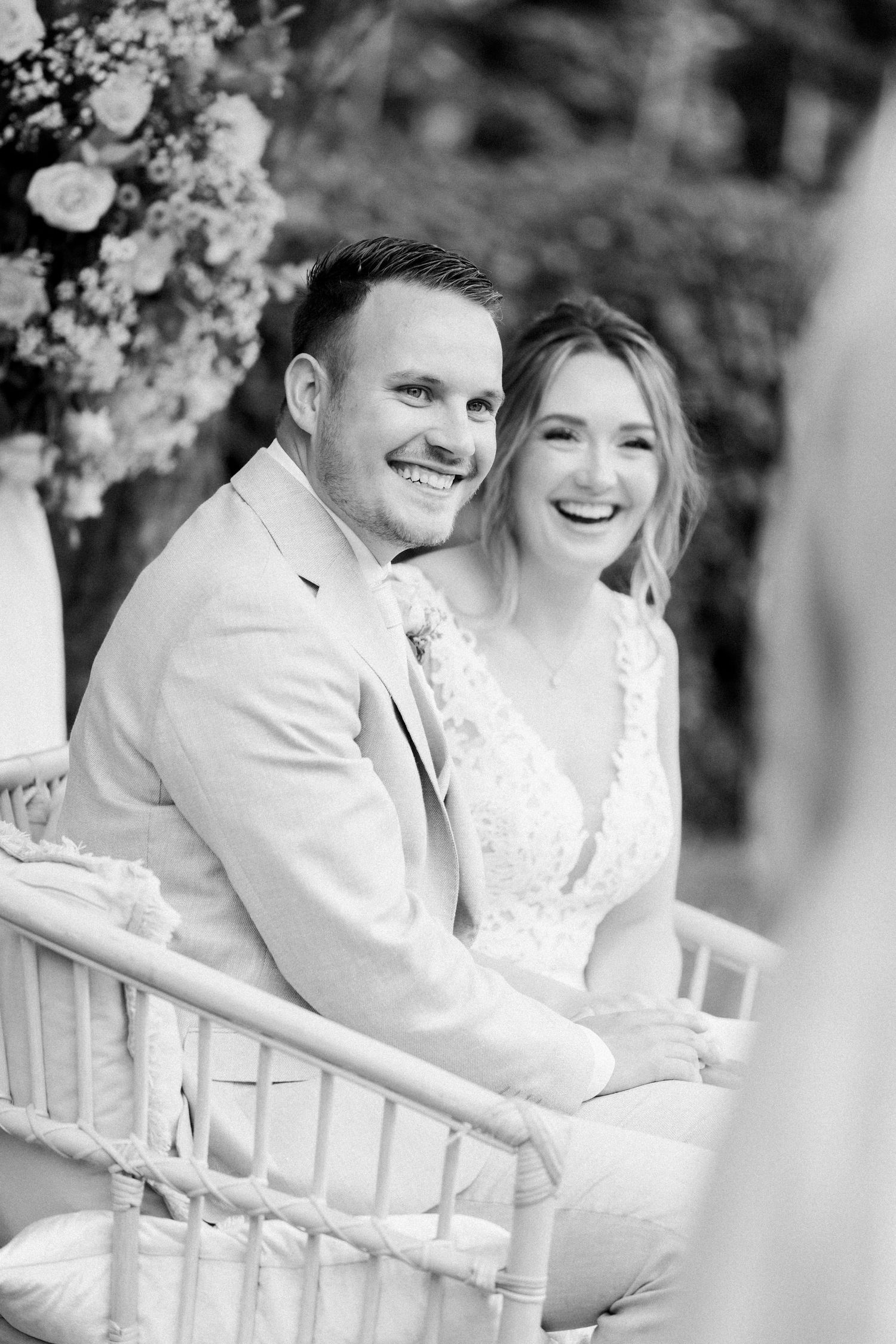 A black and white wedding photo series showing a couple sharing joyful moments while sitting on white chairs outdoors.