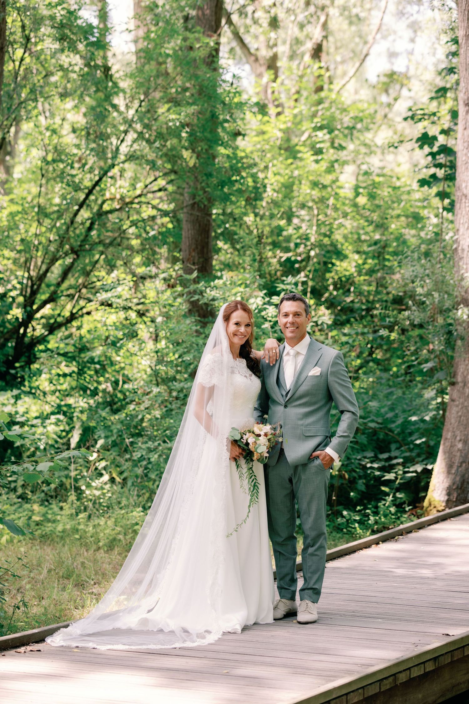 A couple in wedding attire poses together on a wooden boardwalk surrounded by lush green forest scenery.