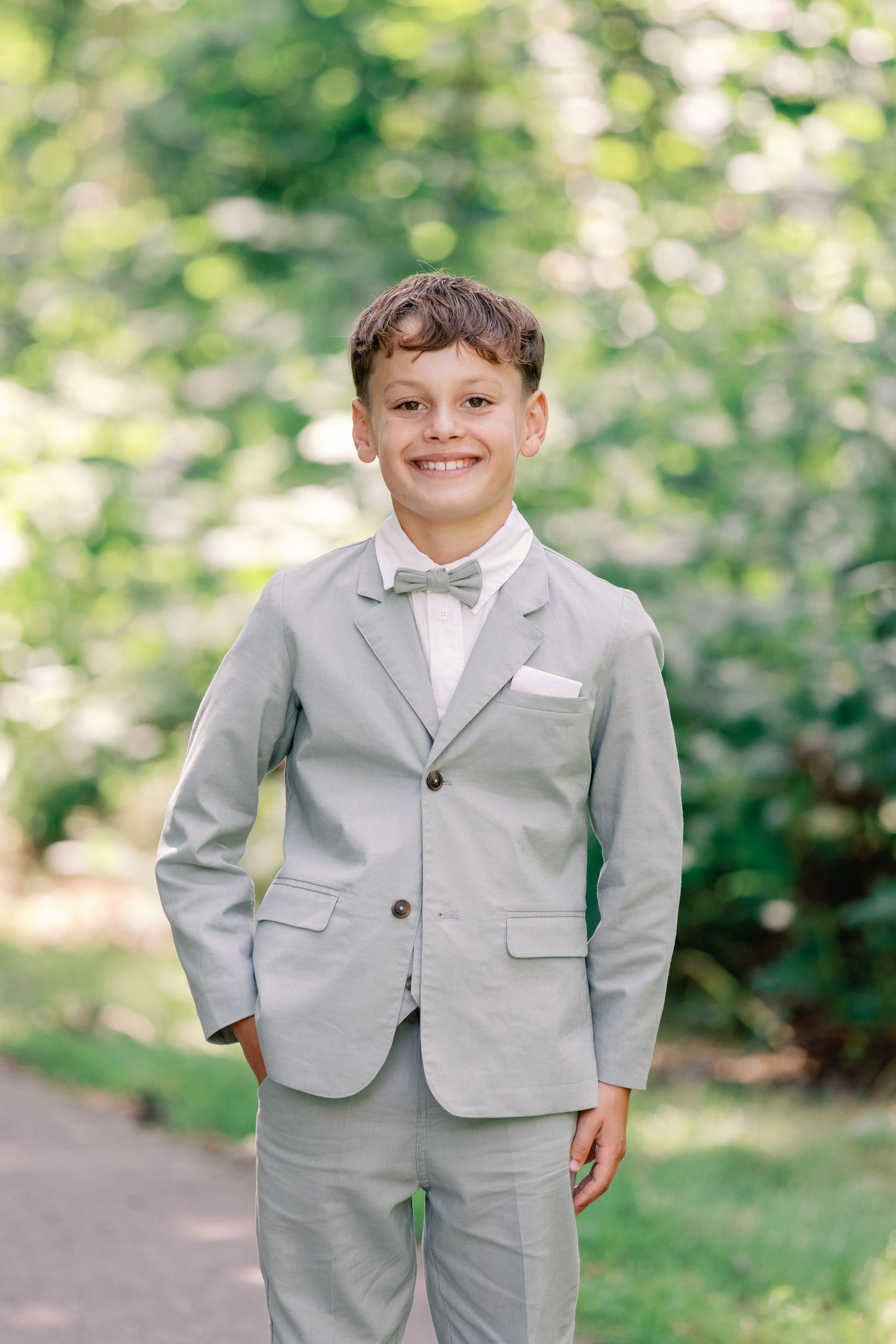 Young ring bearer in a light gray suit poses outdoors against a blurred green garden background.