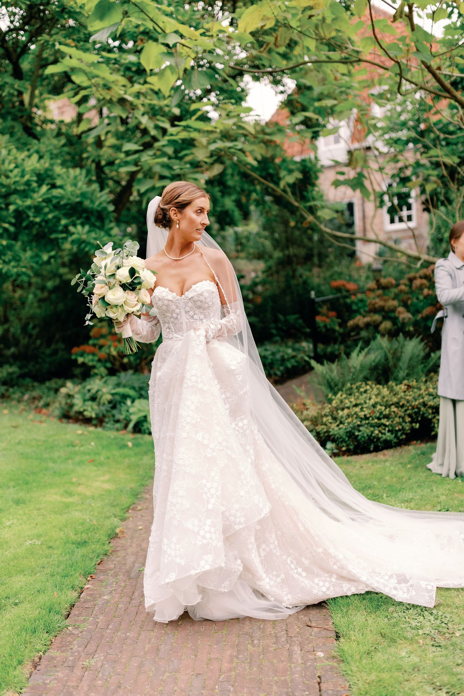 A bride in a flowing white lace gown with a long train poses gracefully in a lush garden setting.