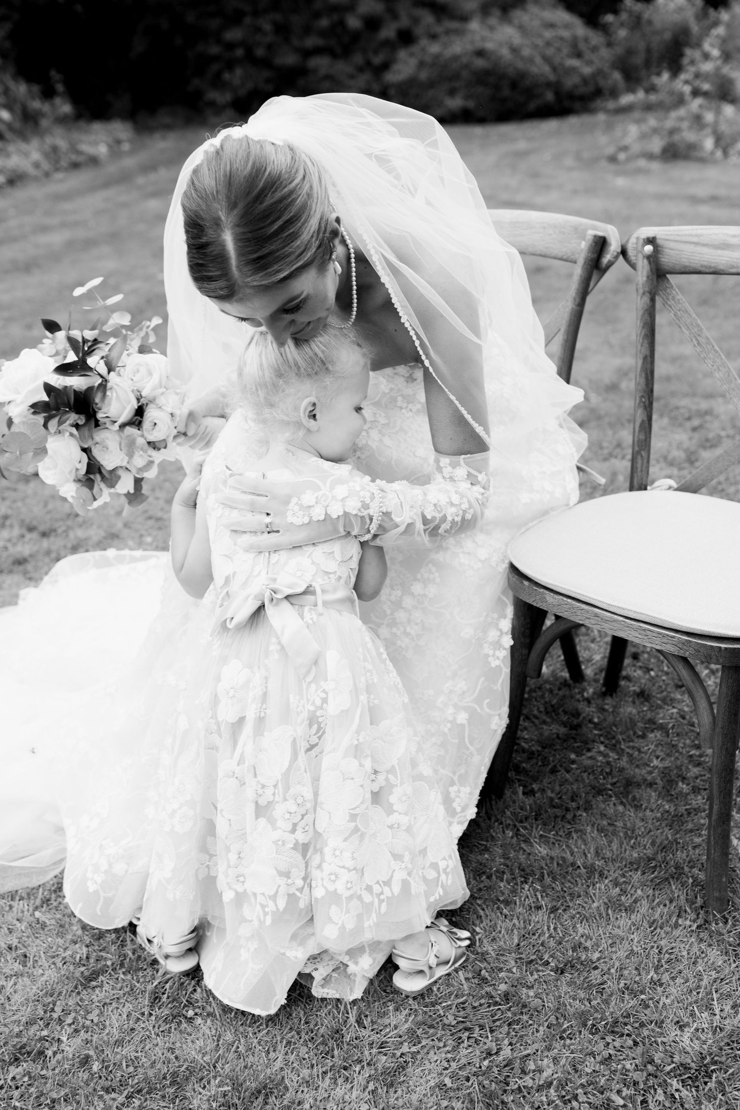 A bride in a flowing white lace wedding dress bends down to pet a small dog on the grass in black and white.