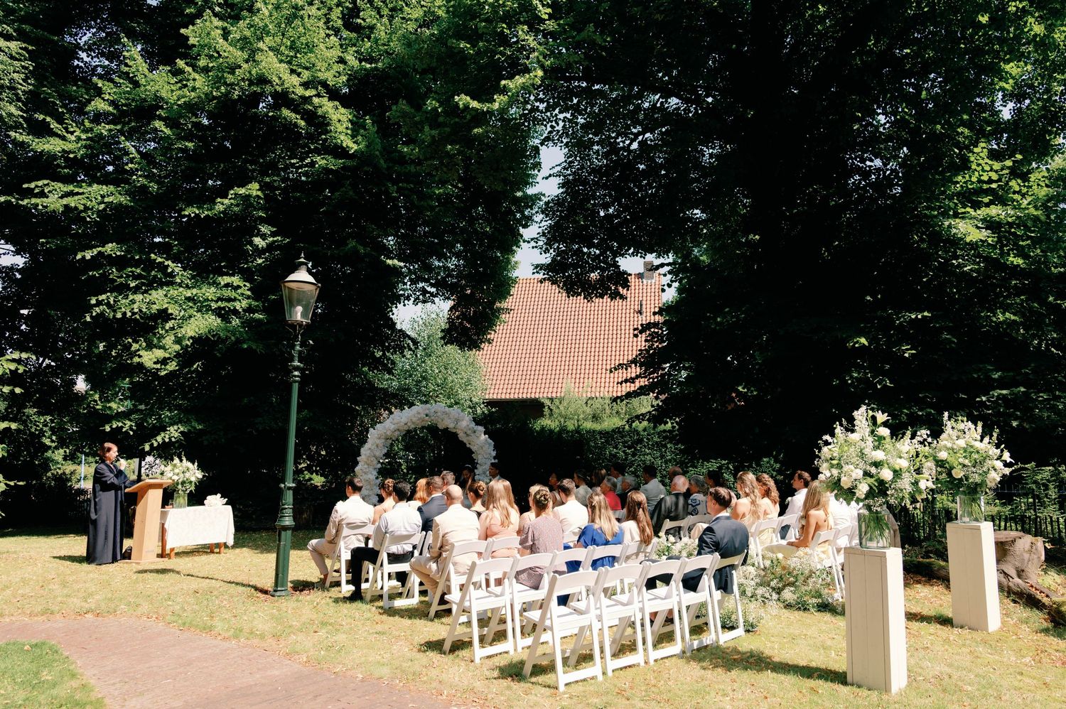 A row of white chairs set up for an outdoor wedding ceremony under tall trees on a sunny day.