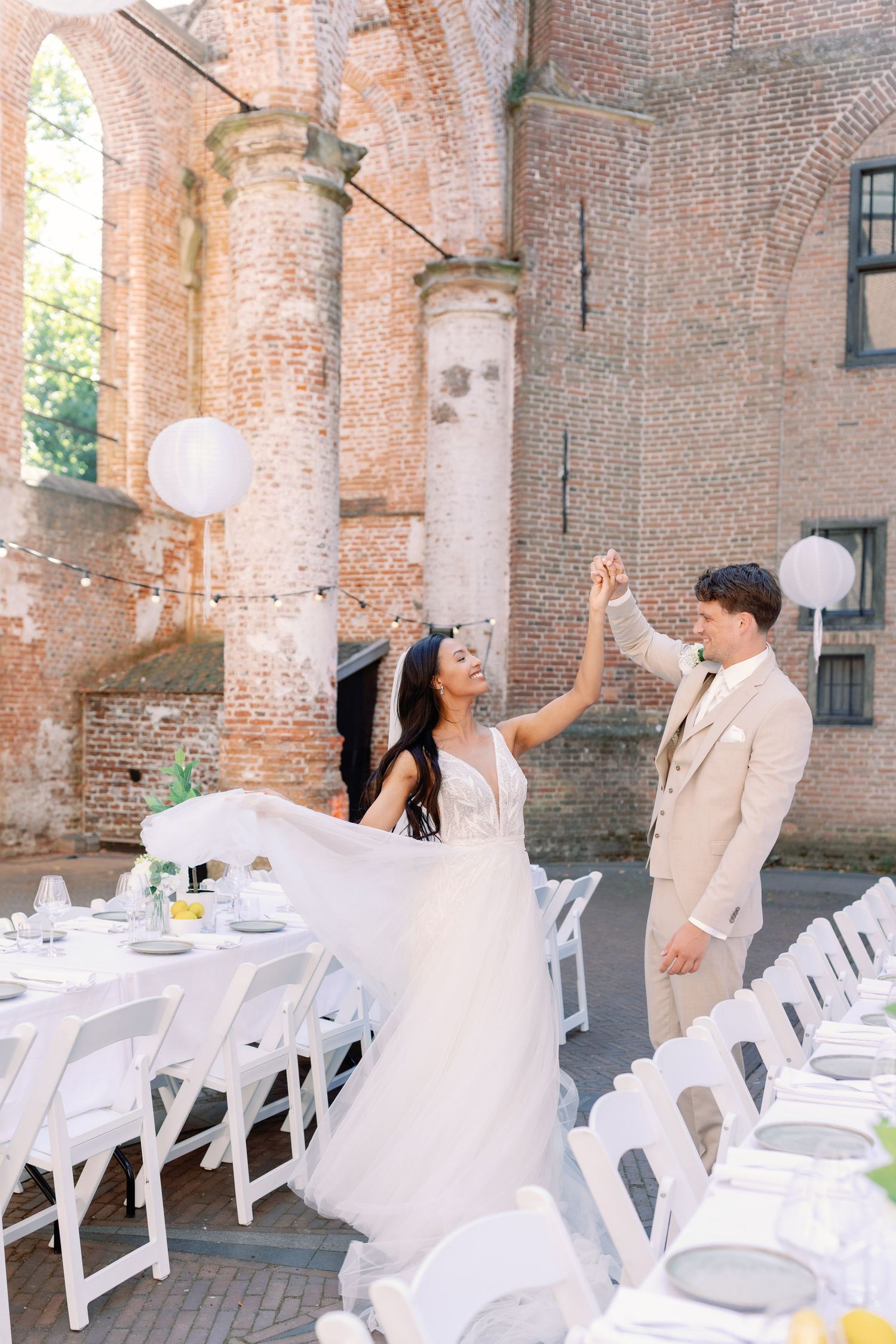 A couple in wedding attire shares a playful moment in a rustic brick venue with white chairs set up for a ceremony.