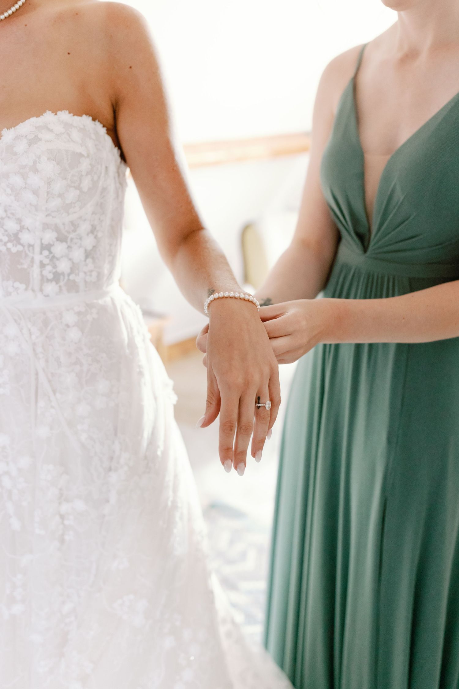 Close-up of bride in lace wedding dress and bridesmaid in emerald green gown holding hands to show jewelry.