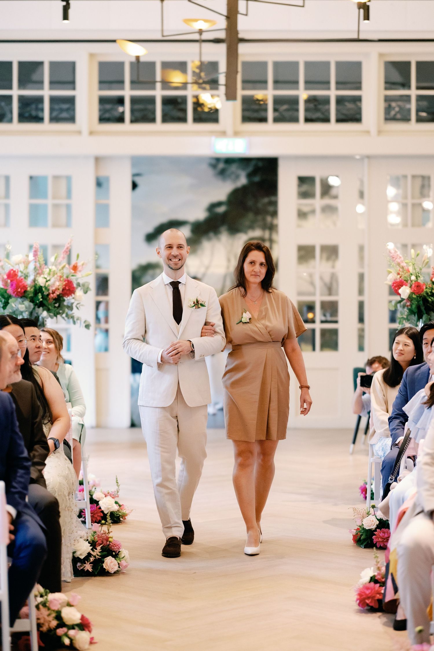 Wedding party members walk down an outdoor corridor lined with pink floral arrangements and seated guests.
