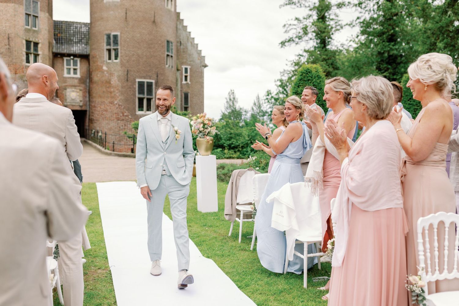 Wedding party members in pink dresses and gray suits celebrate joyfully on a lush green lawn near a stone building.