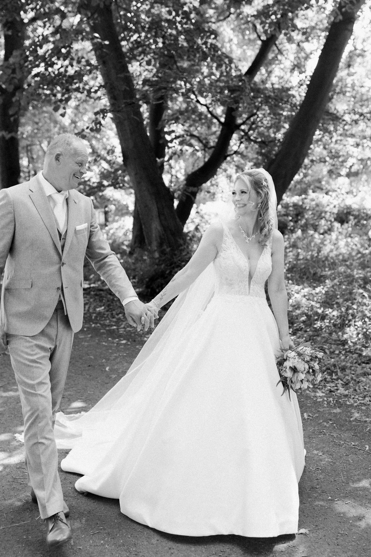 A romantic black and white wedding portrait of a couple walking together under tall trees.
