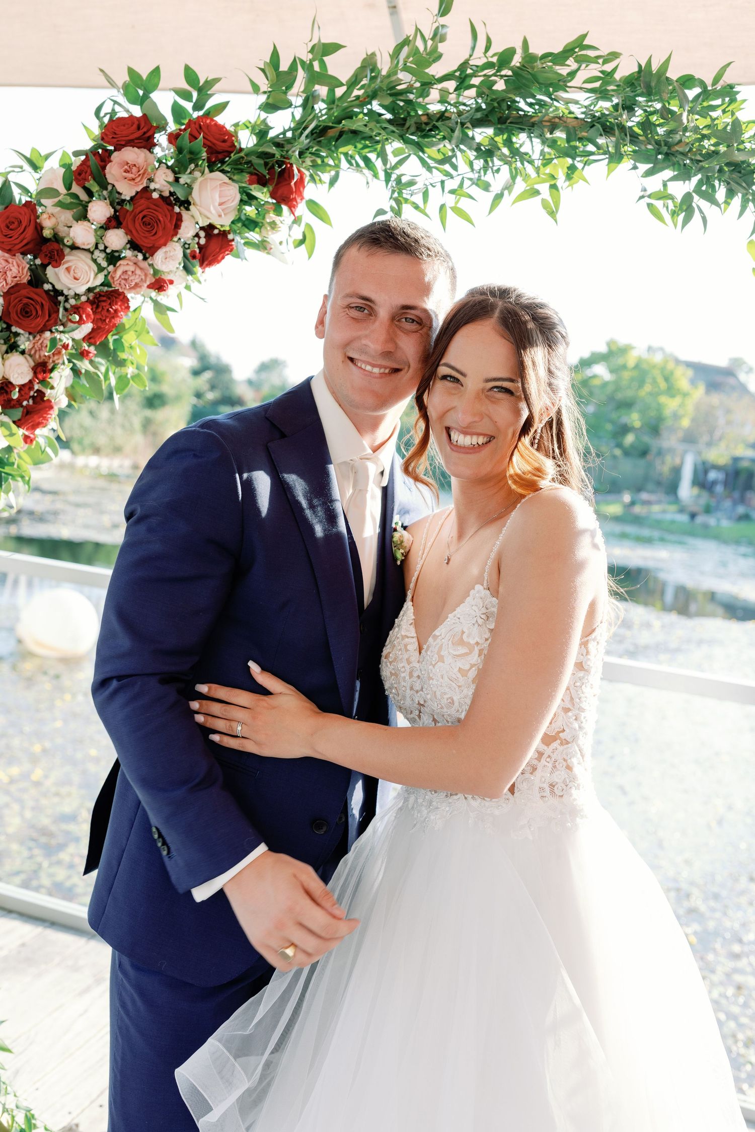 Wedding couple shares intimate moment under floral arch with red roses and greenery.