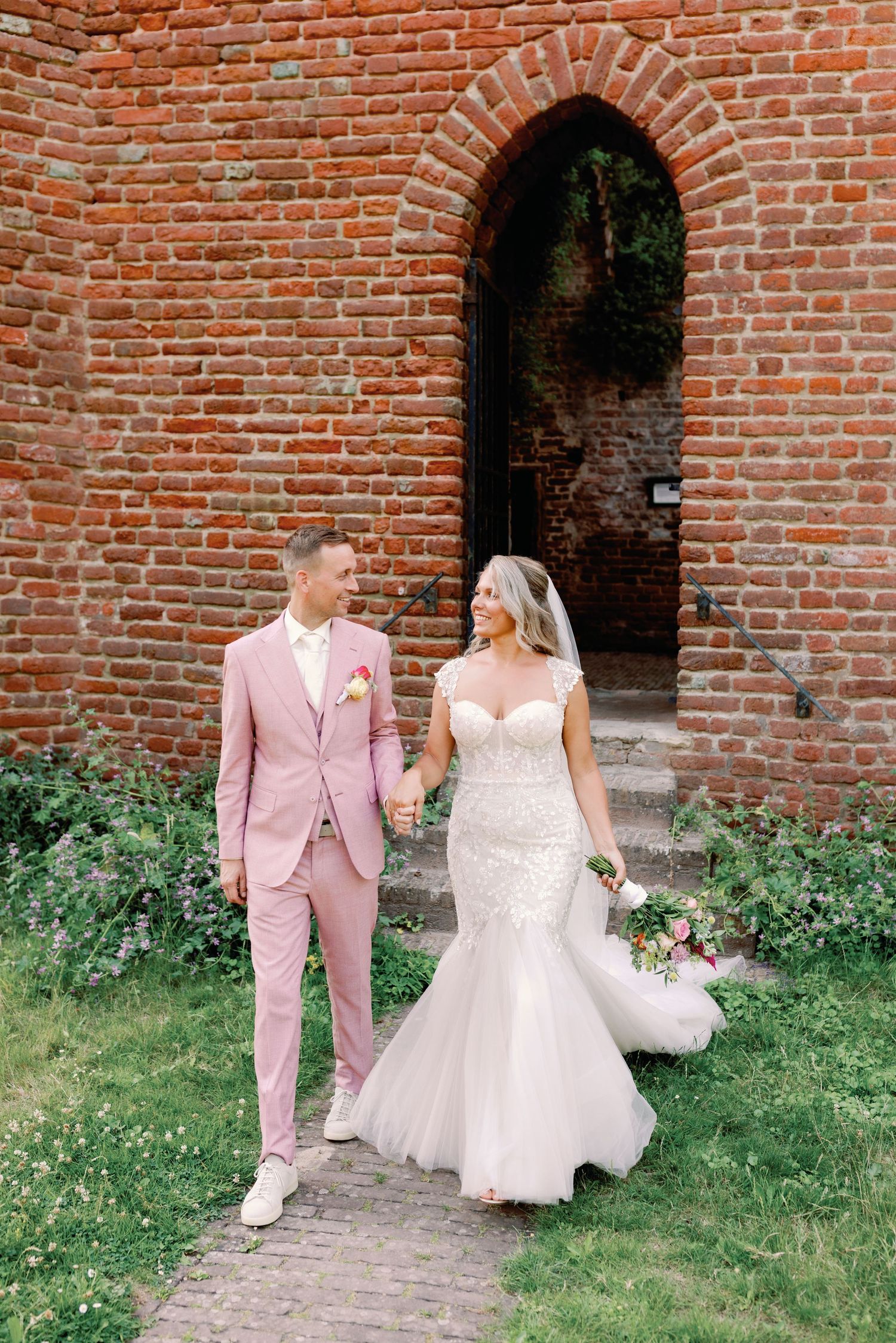 Bride and groom hold hands walking past a red brick archway, she in a lace mermaid gown, he in a pink suit.