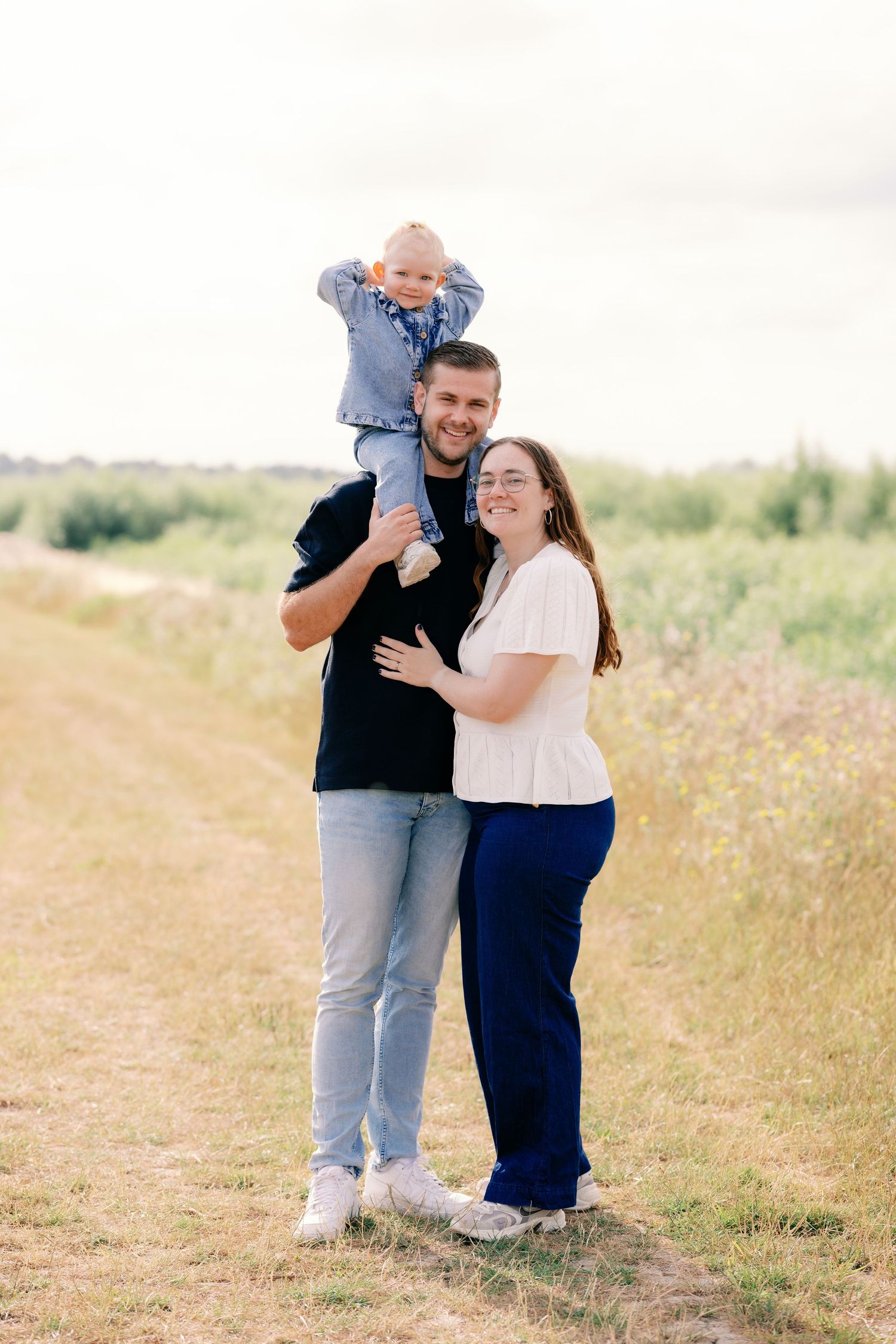 Happy family of three posing outdoors on a dirt path, father holding toddler on shoulders beside smiling mother.
