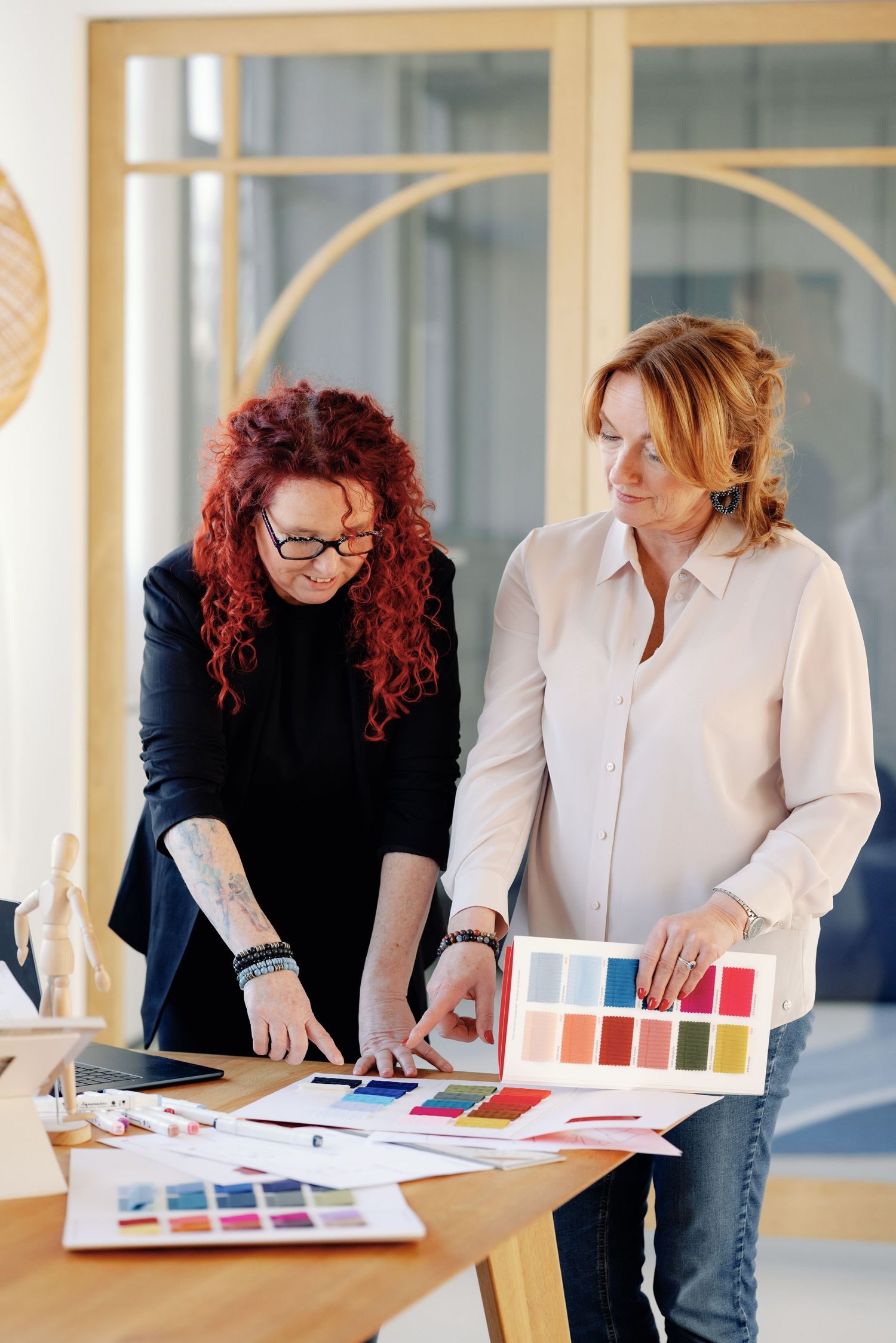 Two designers review color swatches and material samples while collaborating on an interior design project at a wooden table.