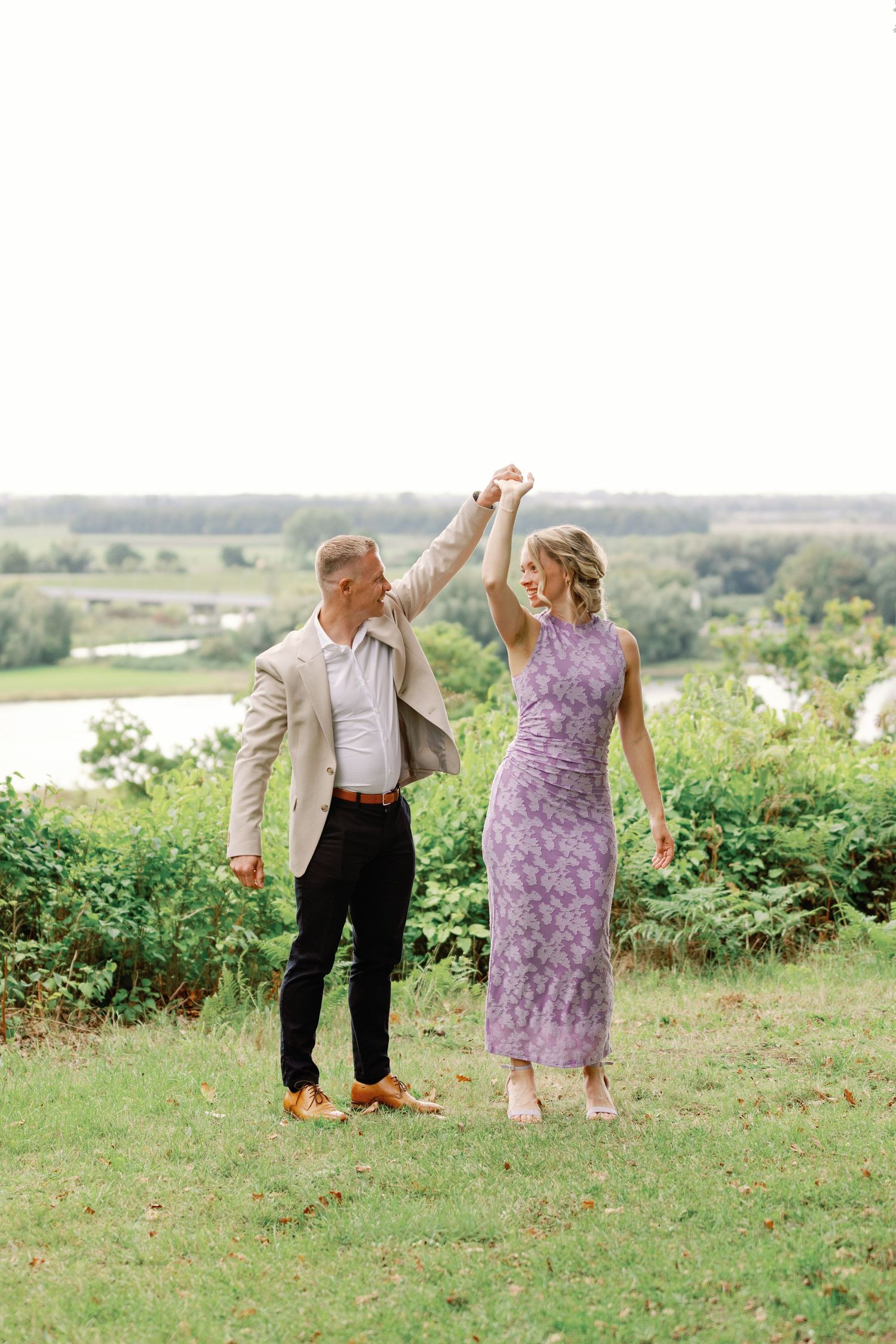 Couple dancing together on a grassy hillside with scenic countryside views in the background.