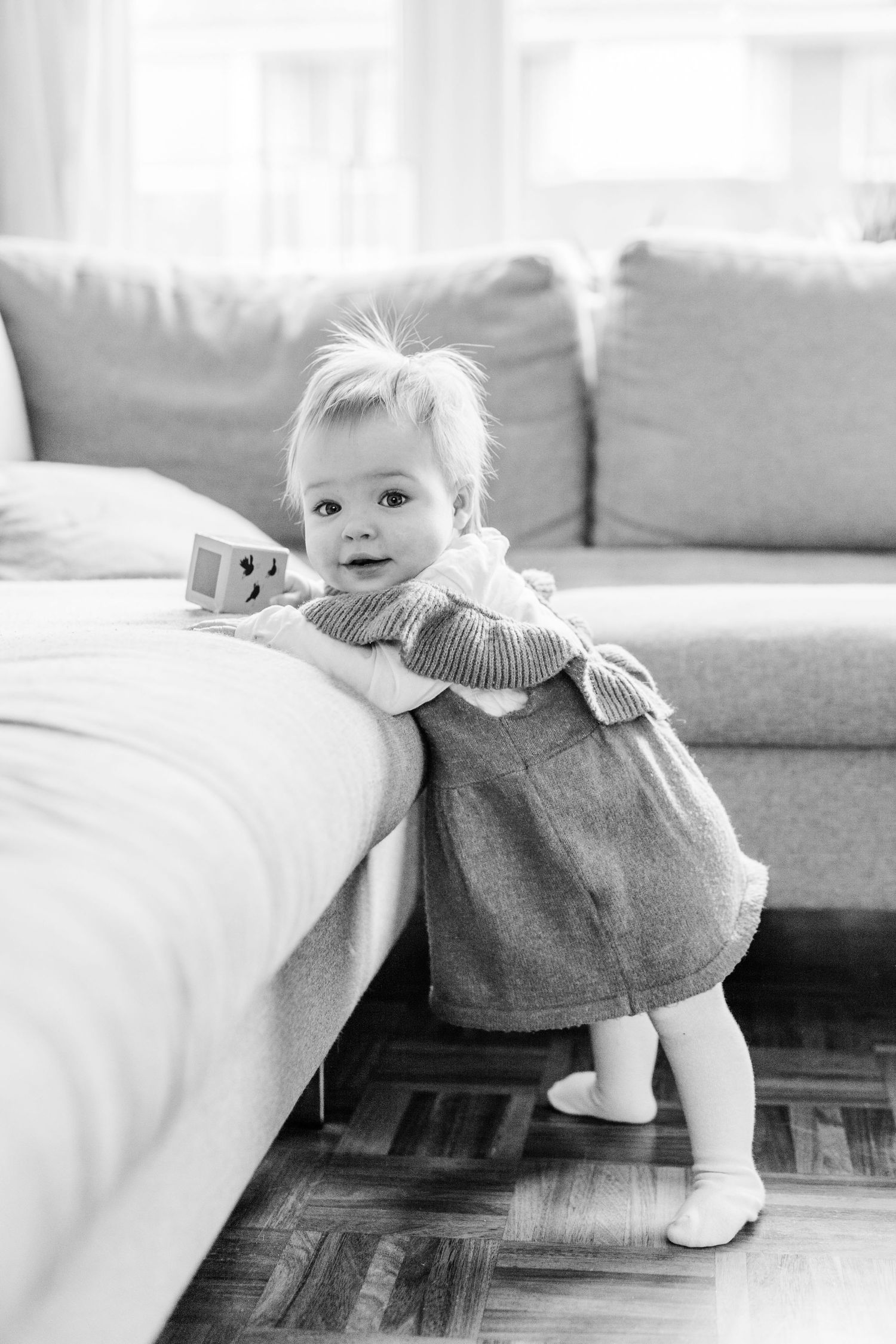 Black and white photo of a toddler in a dress leaning against a sofa in a living room with hardwood floors.